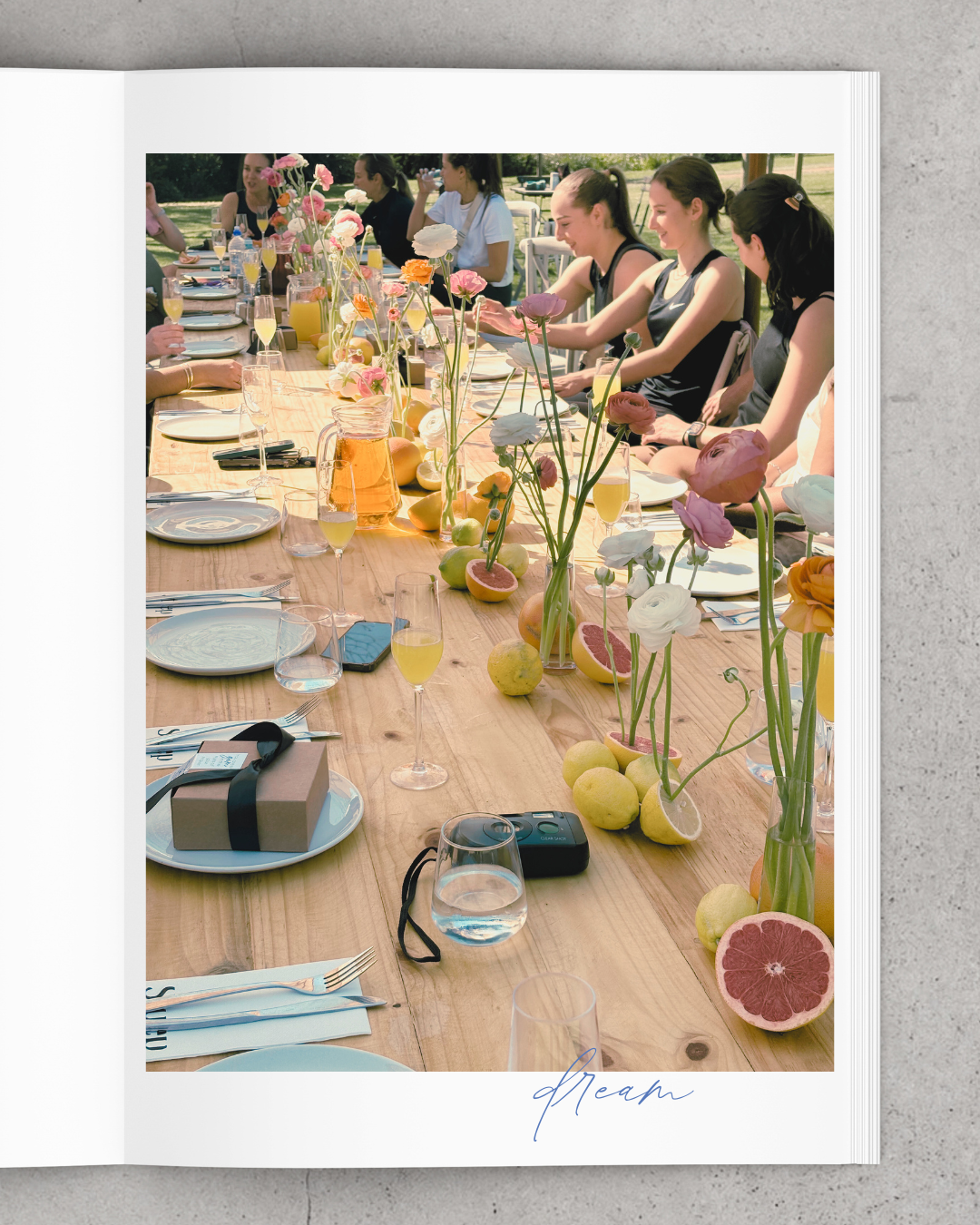 A long outdoor dining table decorated with pink, white, and orange flowers in vases, along with sliced grapefruit, lemons, and apples, set for a gathering with glassware, plates, and cutlery, with women seated and talking in the background.