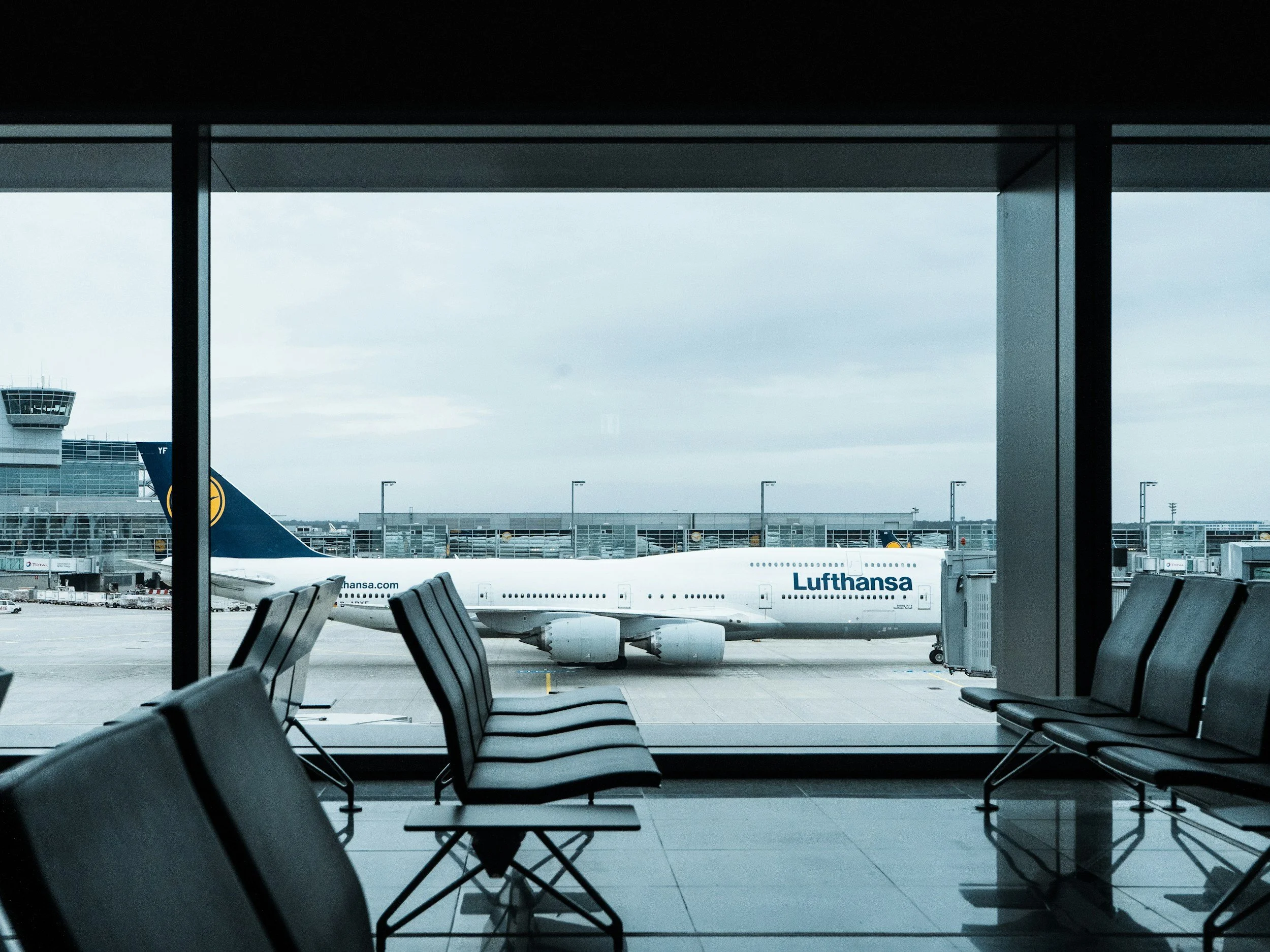 Sitzen im Wartebereich des Flughafens mit Blick auf startendes Lufthansa-Flugzeug im Flug. Der Innenraum ist modern mit schwarzen Sitzen, Blick durch große Fenster auf das Flugfeld.