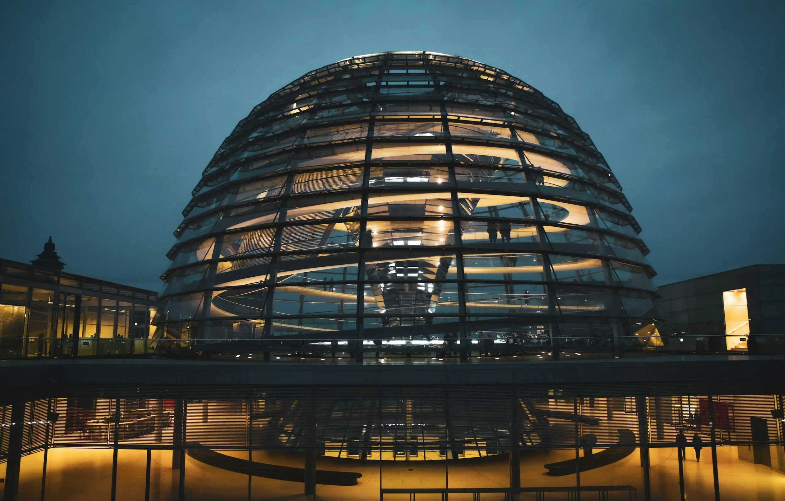 Ein modernes, rundes Glasgebäude bei Nacht, mit sichtbarer spiralförmiger Treppe im Inneren und beleuchteten Etagen, die Menschen zeigen.