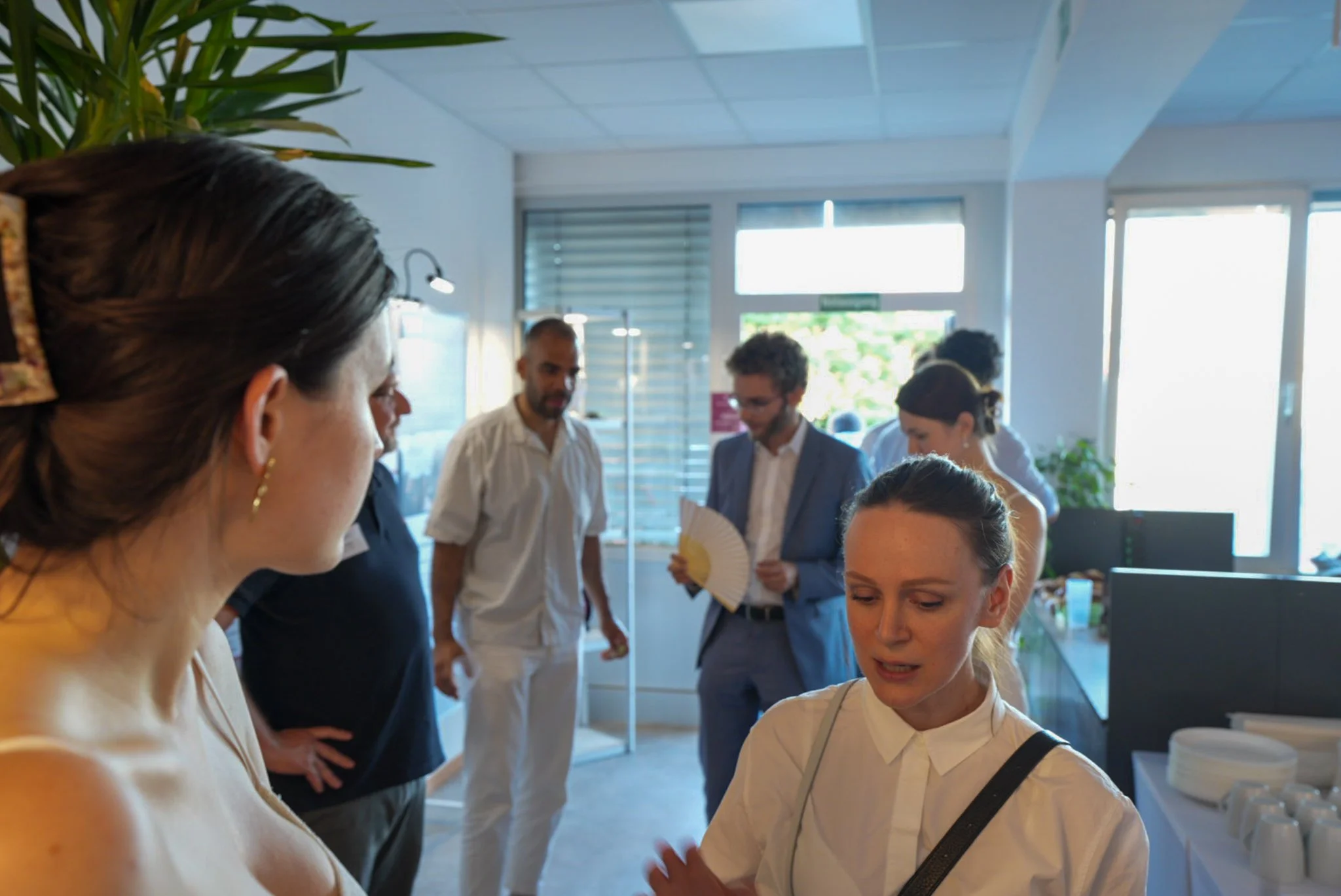 Mehrere Menschen bei einer Veranstaltung im Raum, einige unterhalten sich und lachen, zwei Frauen in der Vordergrund, Hintergrund mit Fenstern und Pflanzen.