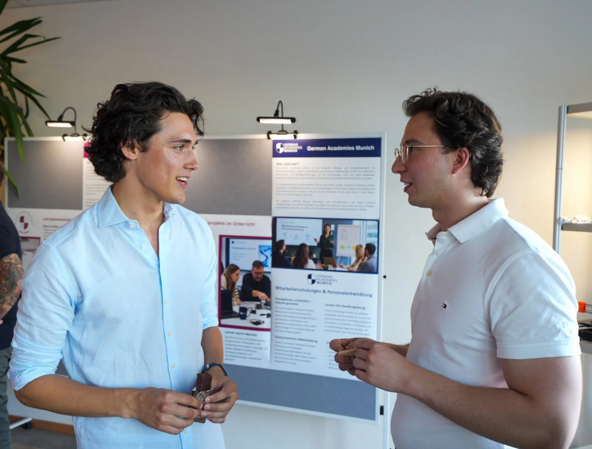 Zwei junge Männer unterhalten sich bei einer Konferenz oder Präsentation, im Hintergrund eine Info-Display mit Text und Bildern.