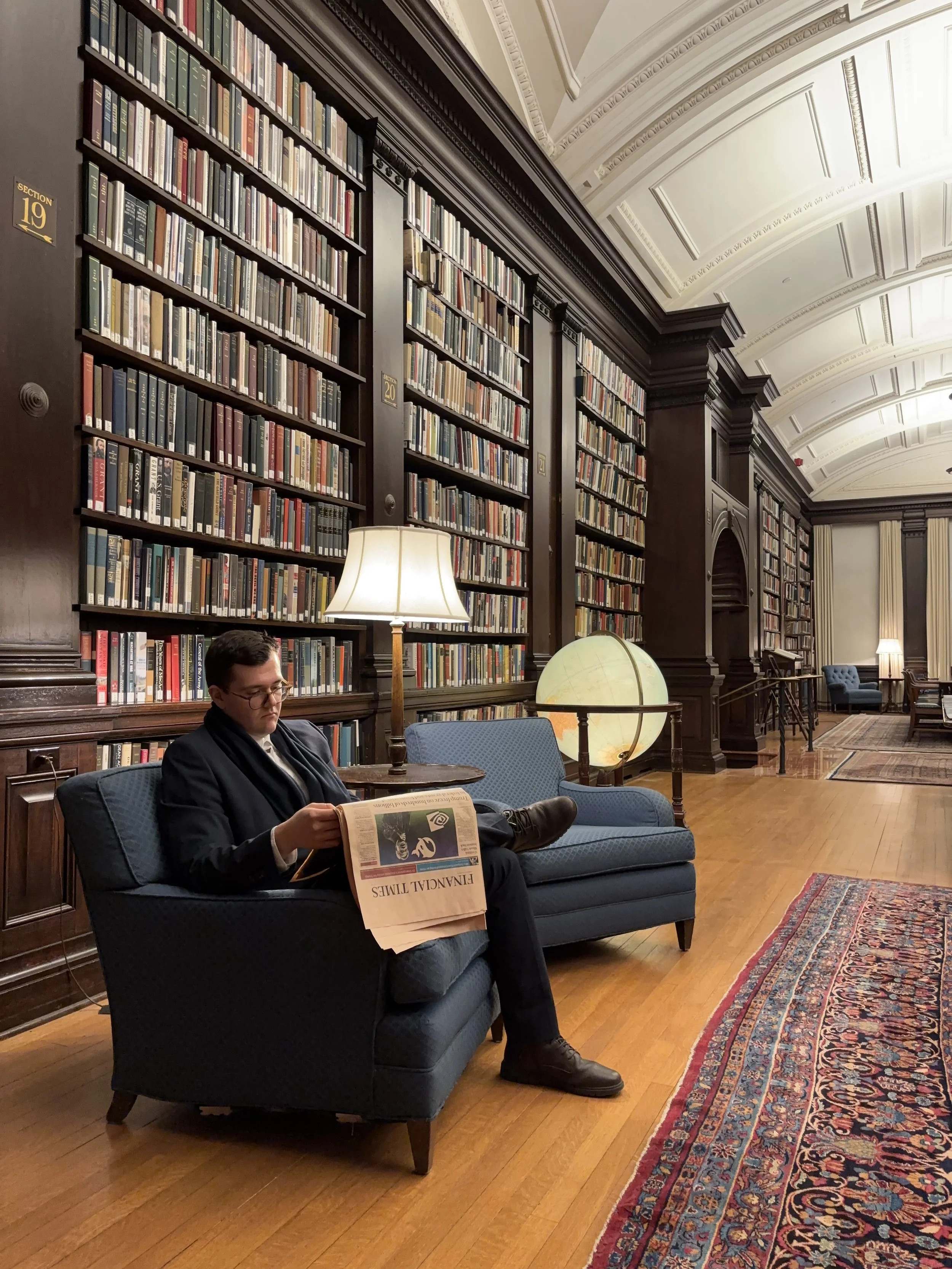 Ein Mann sitzt auf einem blauen Sessel in einer eleganten Bibliothek, liest eine Zeitung namens 'Financial Times', im Hintergrund sind hohe Bücherregale mit vielen Büchern und eine Stehlampe, auf dem Boden liegt ein bunter Teppich.