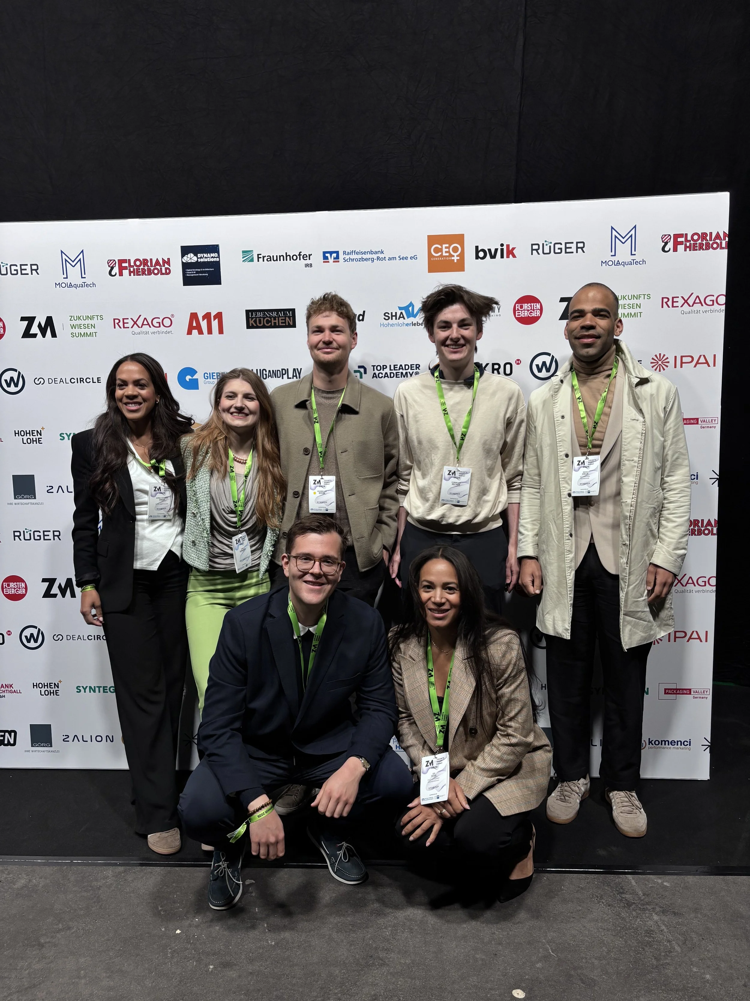 Gruppenfoto von neun jungen Erwachsenen, die auf einer Konferenz mit schwarzen Hintergrund und einer großen Sponsorenwand stehen. Die Teilnehmer tragen Namensschilder und Lanyards und lächeln in die Kamera.