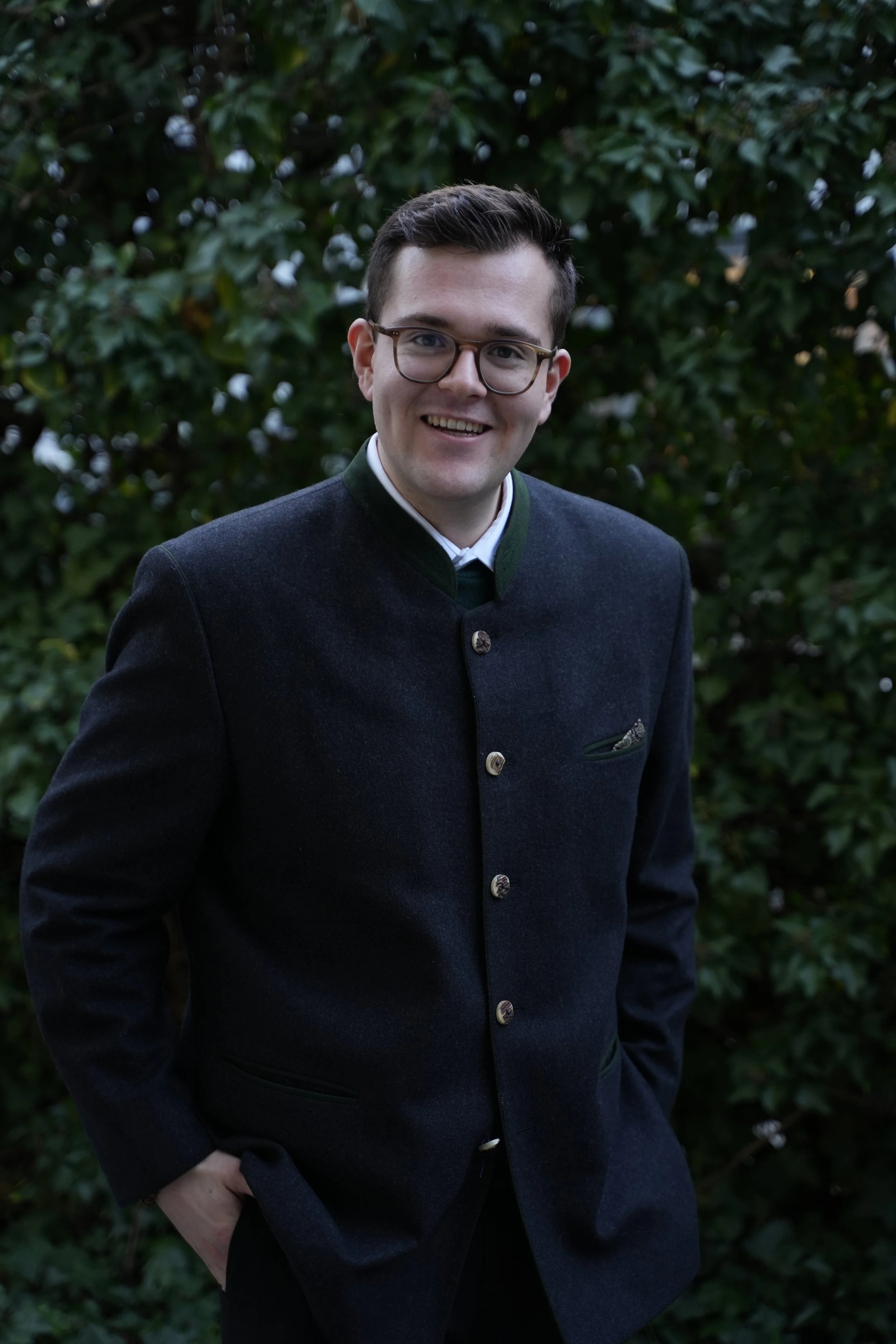 Ein junger Mann trägt eine traditionelle dunkle Jacke mit Knöpfen und Brille, steht vor einer Büschel im Freien und lächelt in die Kamera.