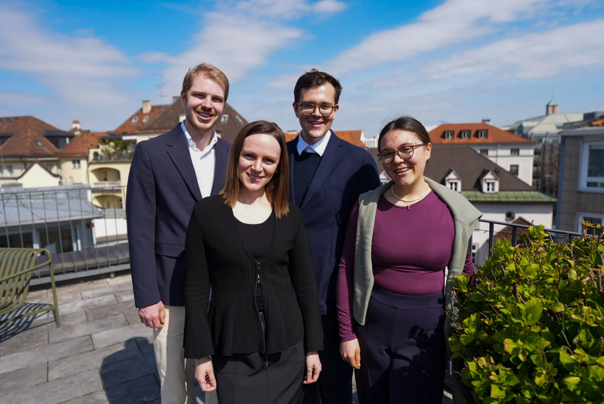 Fünf Menschen auf einem Dach mit Stadtblick und blauen Himmel im Hintergrund, lachend und in formeller Kleidung.