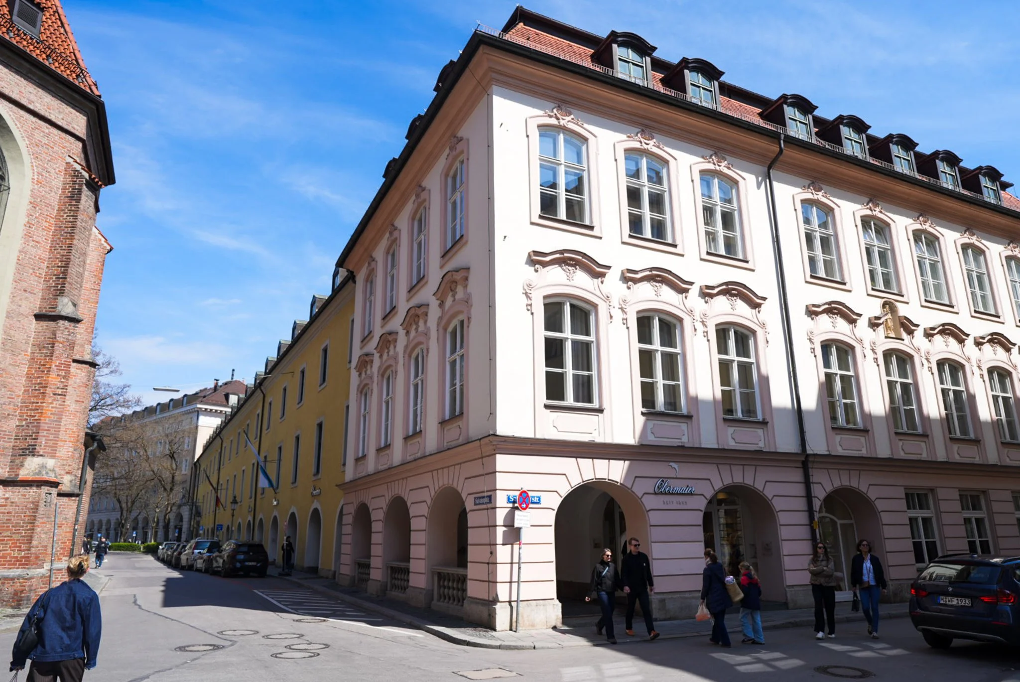 Mehrstöckiges Gebäude mit barocken Fassadendetails, große Fenster und einem roten Ziegeldach, in einer Stadtstraße mit Passanten und parkenden Autos.
