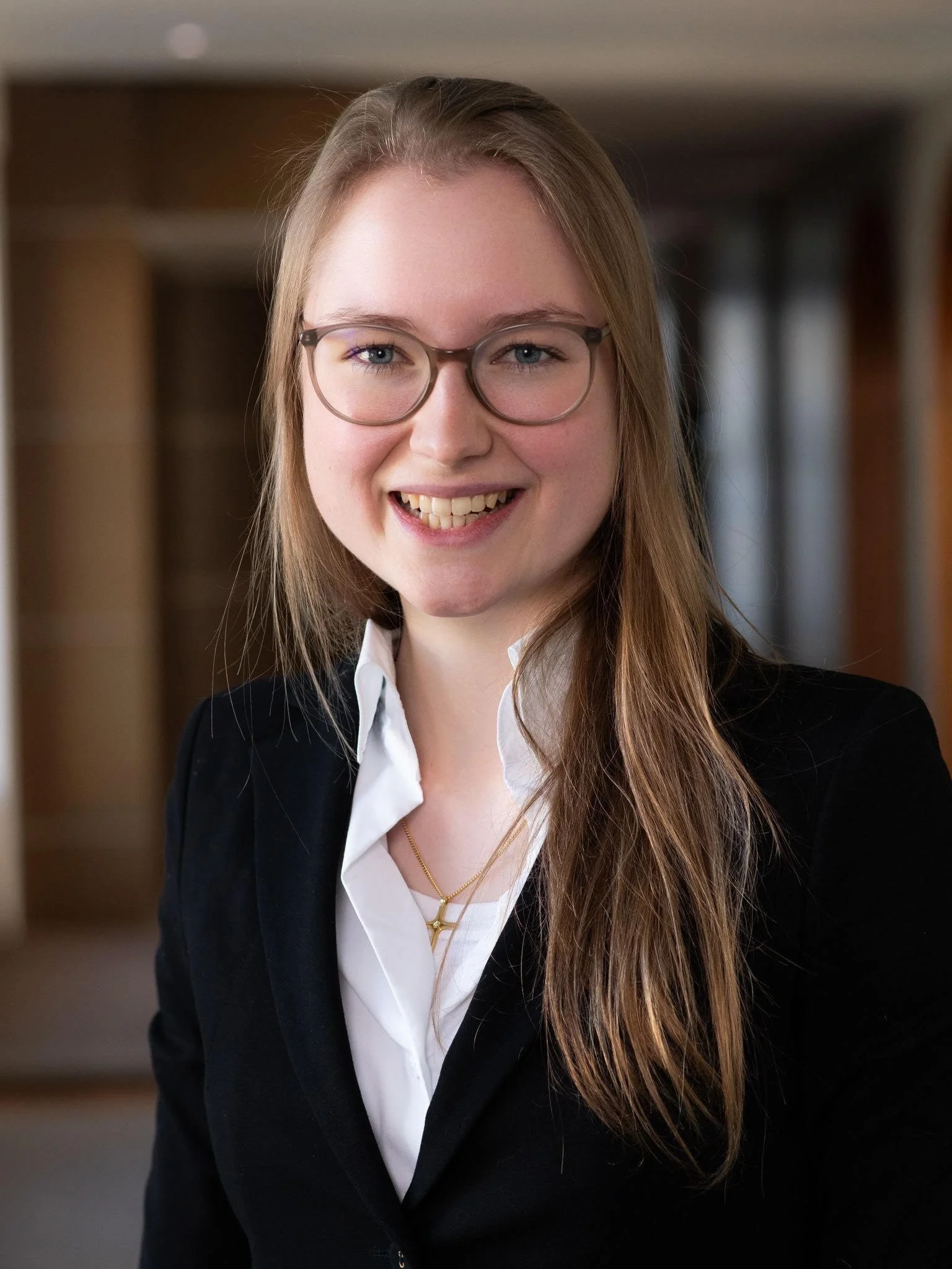 Junge Frau mit langen braunen Haaren, Brille, weiße Bluse und schwarzem Blazer, lächelnd, vor unscharfem Hintergrund.
