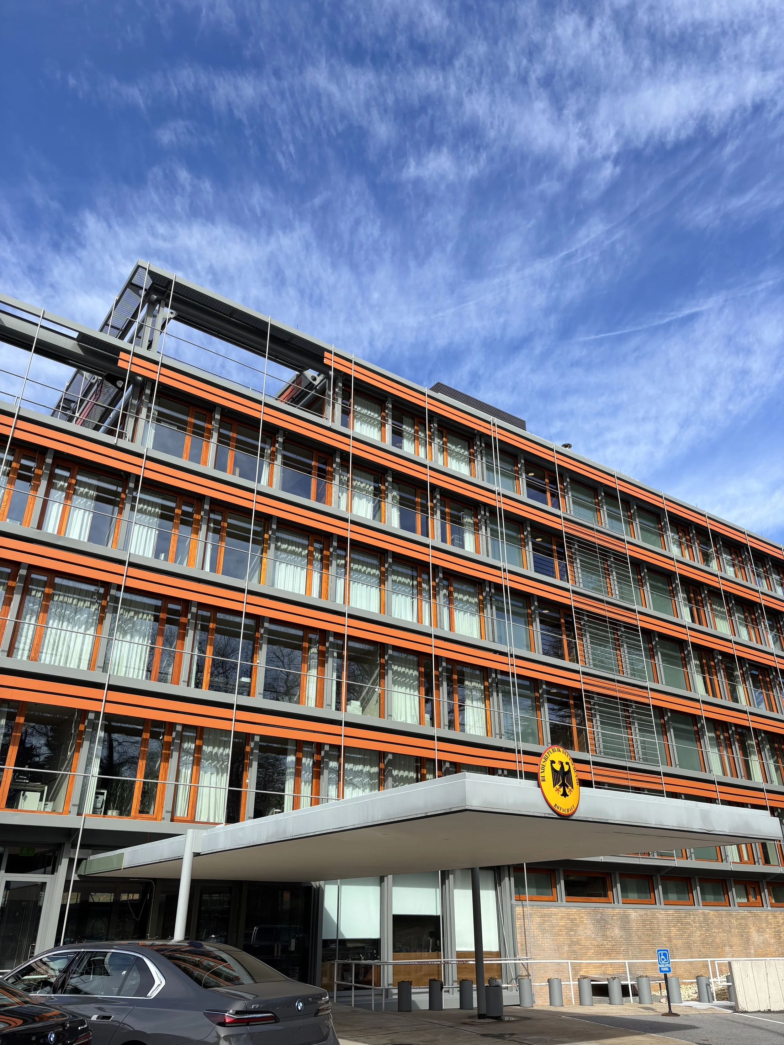 Ein modernes Bürogebäude mit mehreren Stockwerken, großen Fenstern und einem deutschen Wappen an der Fassade. Es gibt Parkplätze und eine Behindertentoilette im Vordergrund.