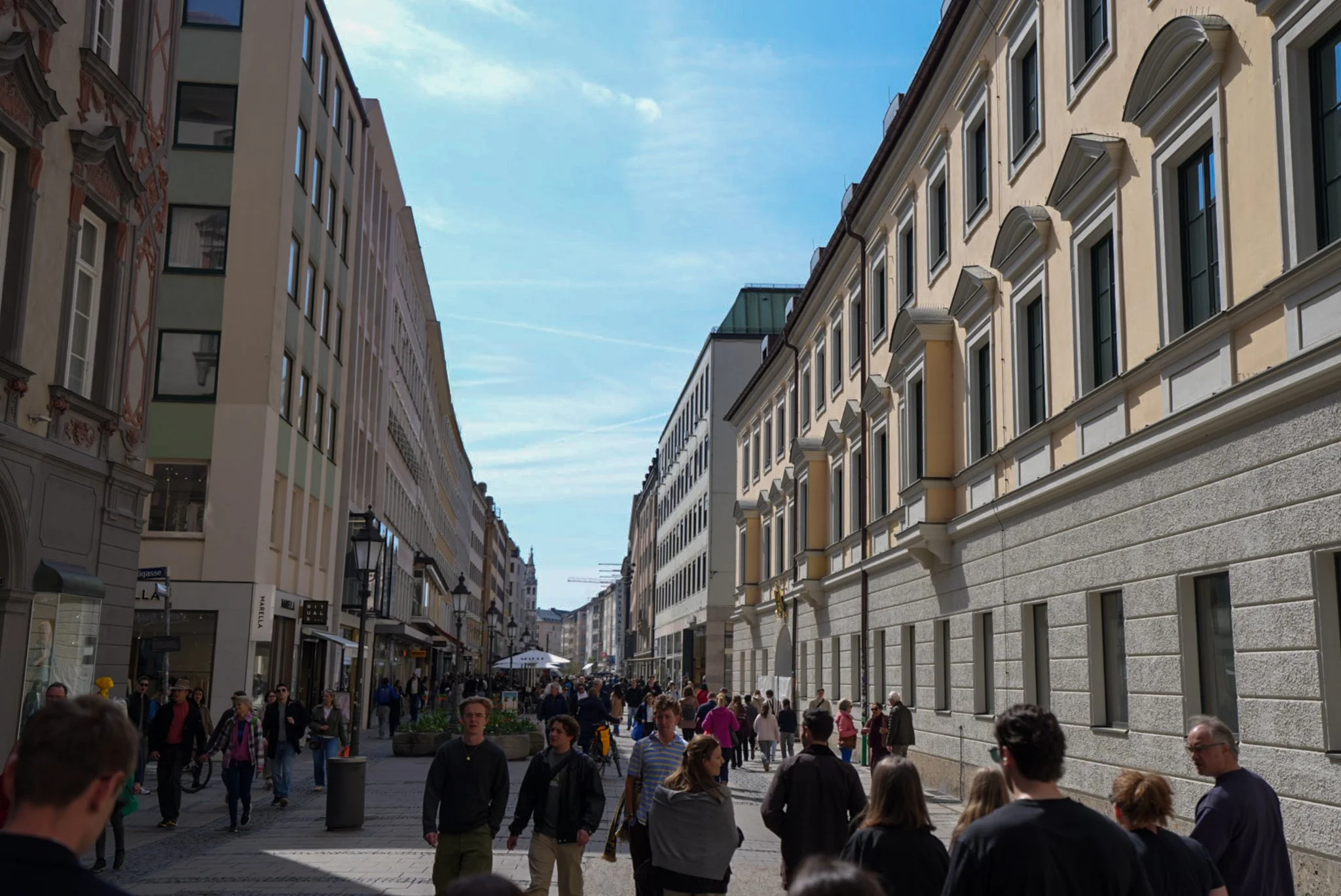 Eine geschäftige Straßenszene in einer europäischen Stadt mit vielen Passanten, umgeben von historischen Gebäuden mit großen Fenstern und dekorativen Fassaden, bei schönem Wetter mit blauer Himmel.