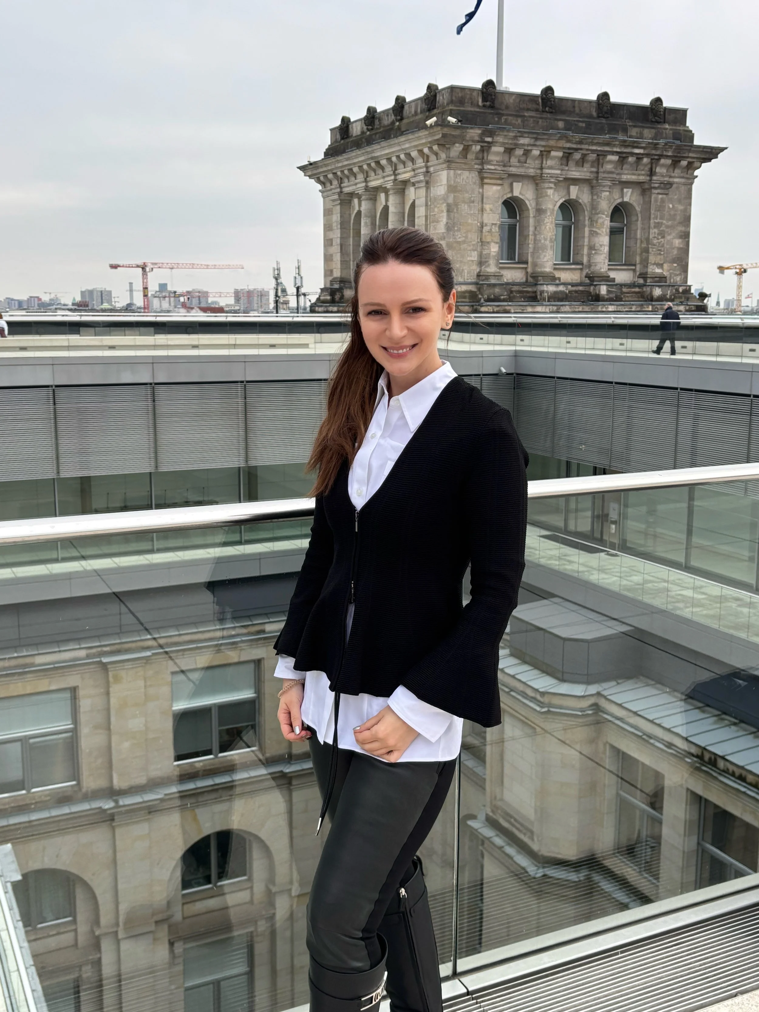 Junge Frau mit braunen Haaren, weißem Hemd, in einem modernen Büro mit großen Fenstern im Hintergrund.
