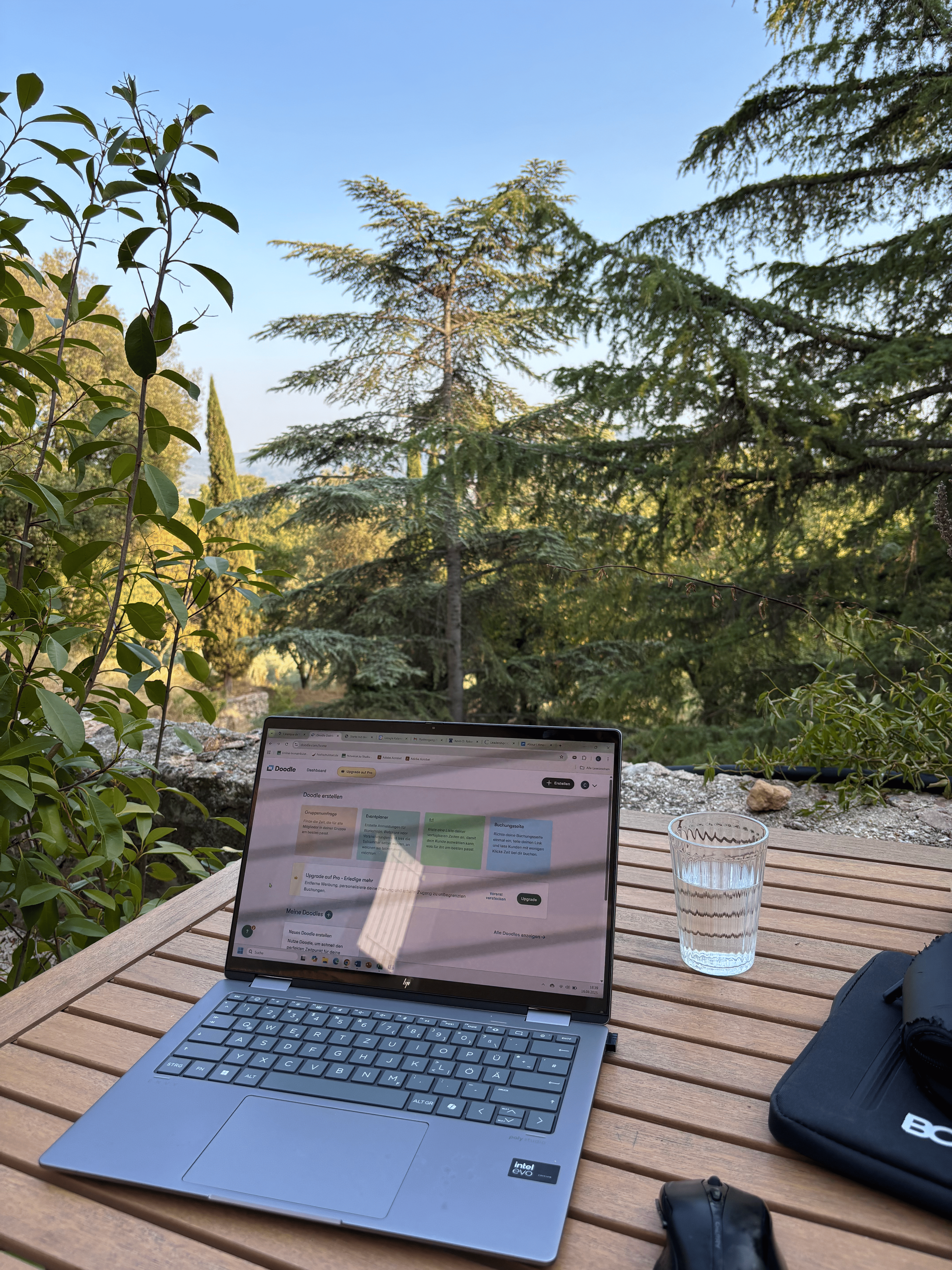 Laptop auf einem Holztisch im Freien mit Blick auf Bäume und blauen Himmel, daneben ein Wasserglas und eine schwarze Tasche.