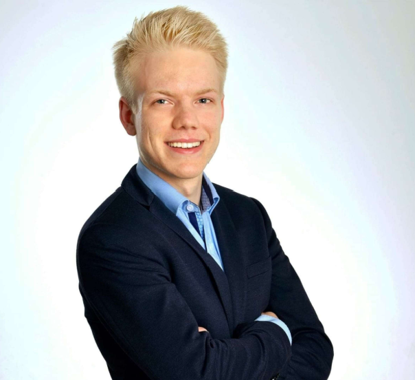 Ein junger Mann mit blonden Haaren, blauer Hemd und dunklem Blazer lächelnd mit verschränkten Armen vor hellem Hintergrund.