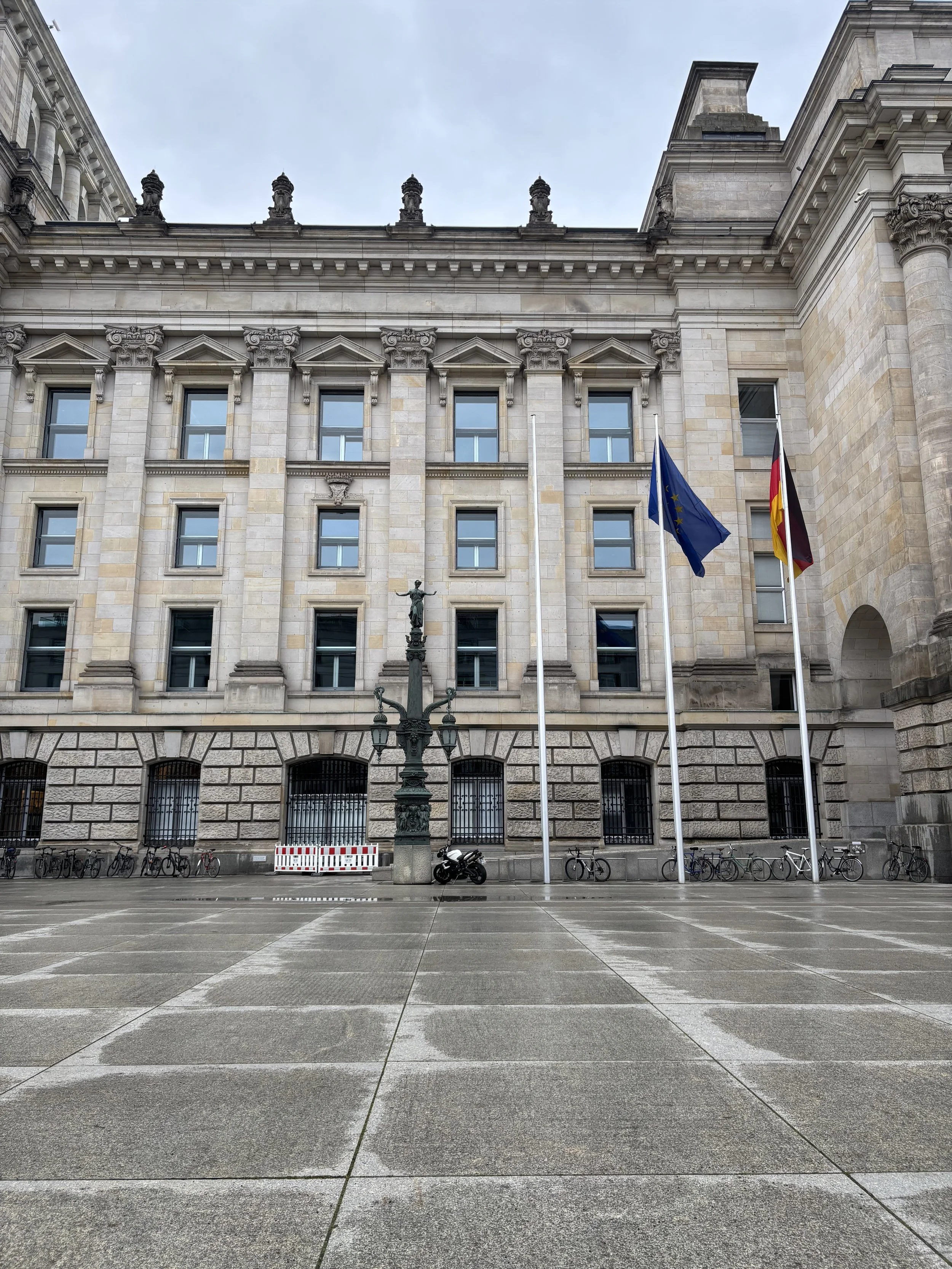 Ein modernes Geschäftsgebäude mit mehreren Etagen und großen Fenstern. Es gibt ein Schild mit dem deutschen Bundesadler und dem Wort 'Deutschland'. Im Vordergrund sind Autos auf einem Parkplatz sichtbar, darunter ein schwarzer BMW. Es ist ein blauer Himmel mit einigen Wolken.
