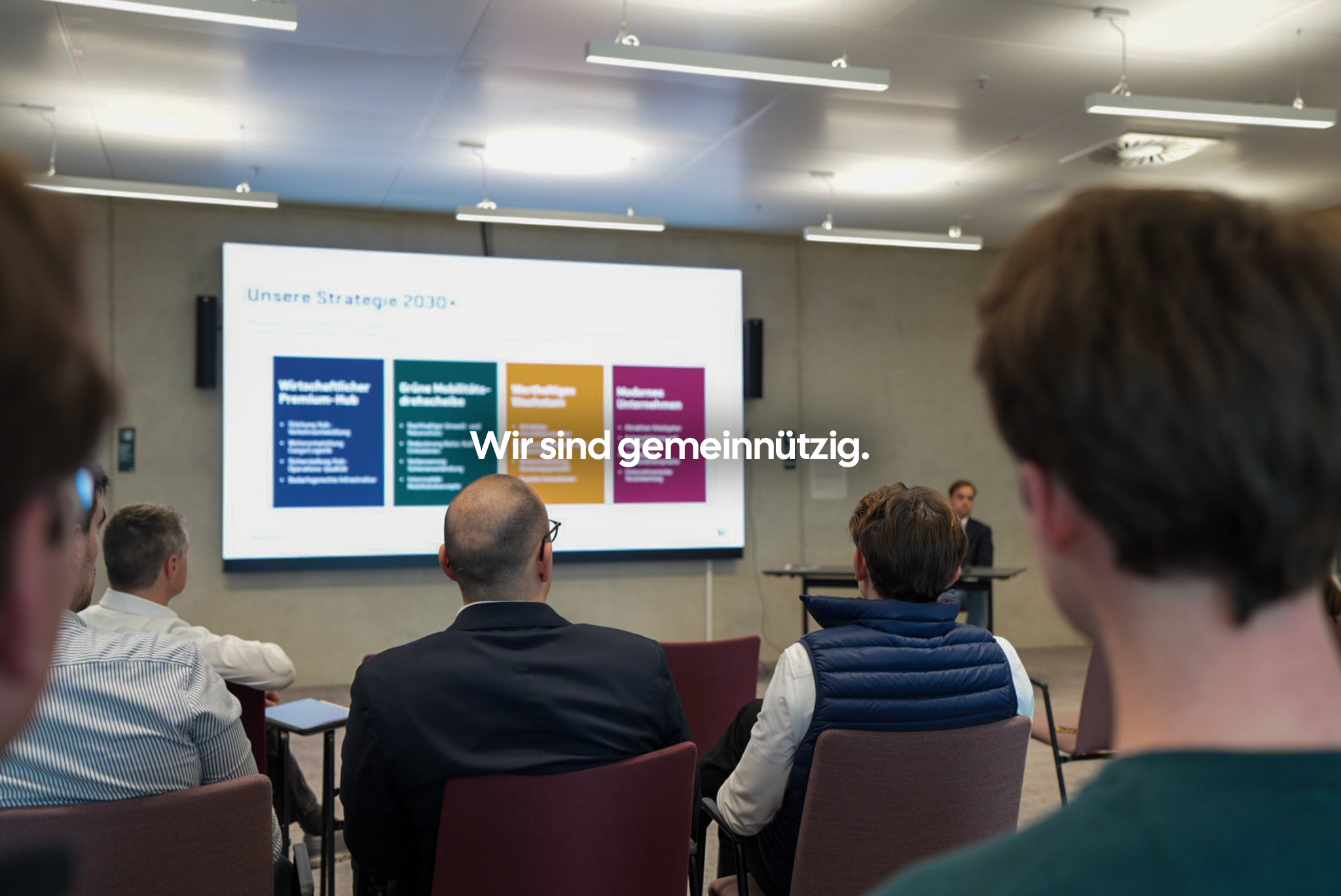 Innenaufnahme eines Konferenzraums mit mehreren sitzenden Personen, die einem Präsentationsbild an der Wand folgen. Das Display zeigt eine Strategiepräsentation mit Überschrift "Unsere Strategie 2030" und vier farbige Abschnitte.
