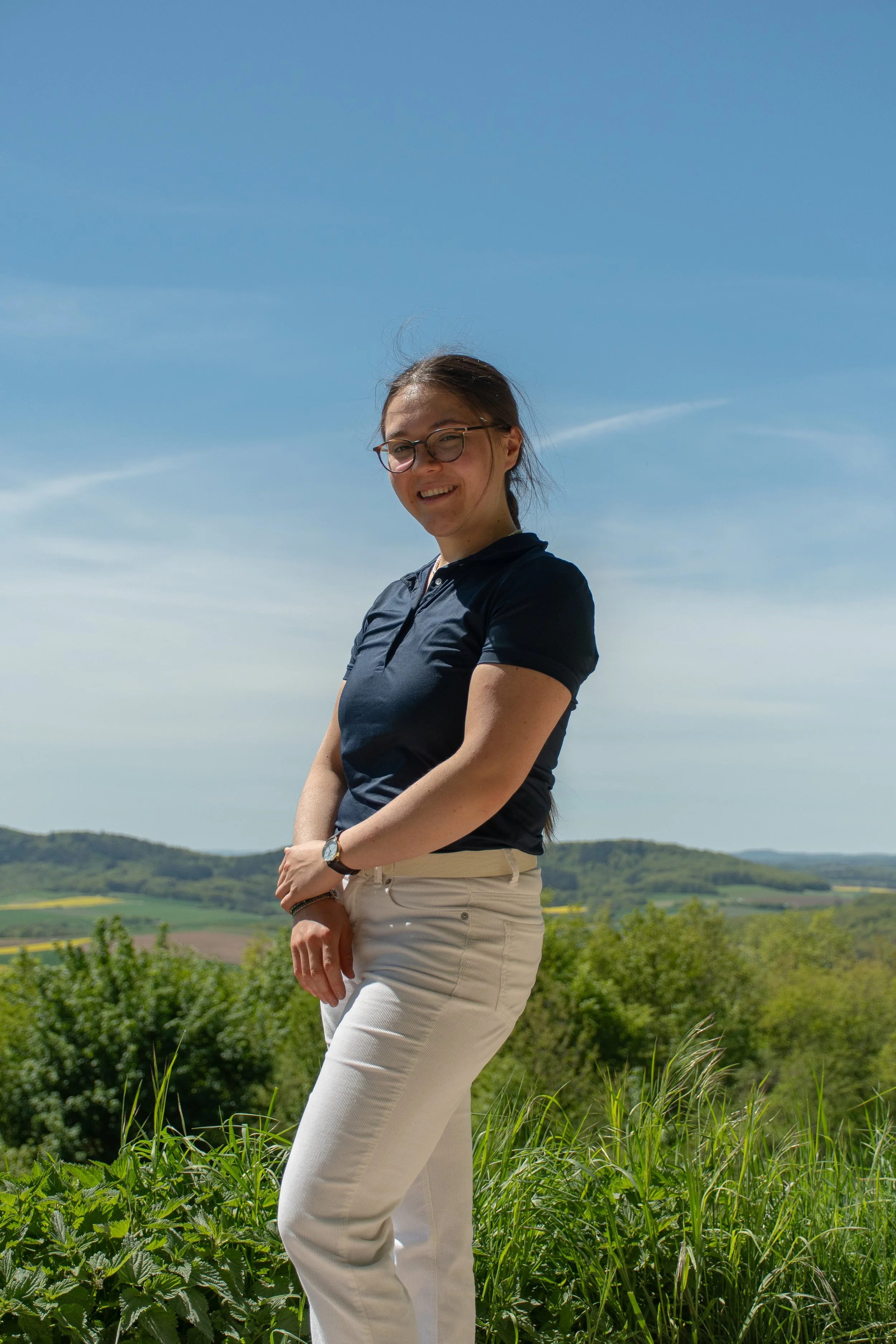 Eine lachende Frau mit Brille, schwarzen Haaren und hellen Hosen im Freien vor einer grünen Landschaft unter blauem Himmel.