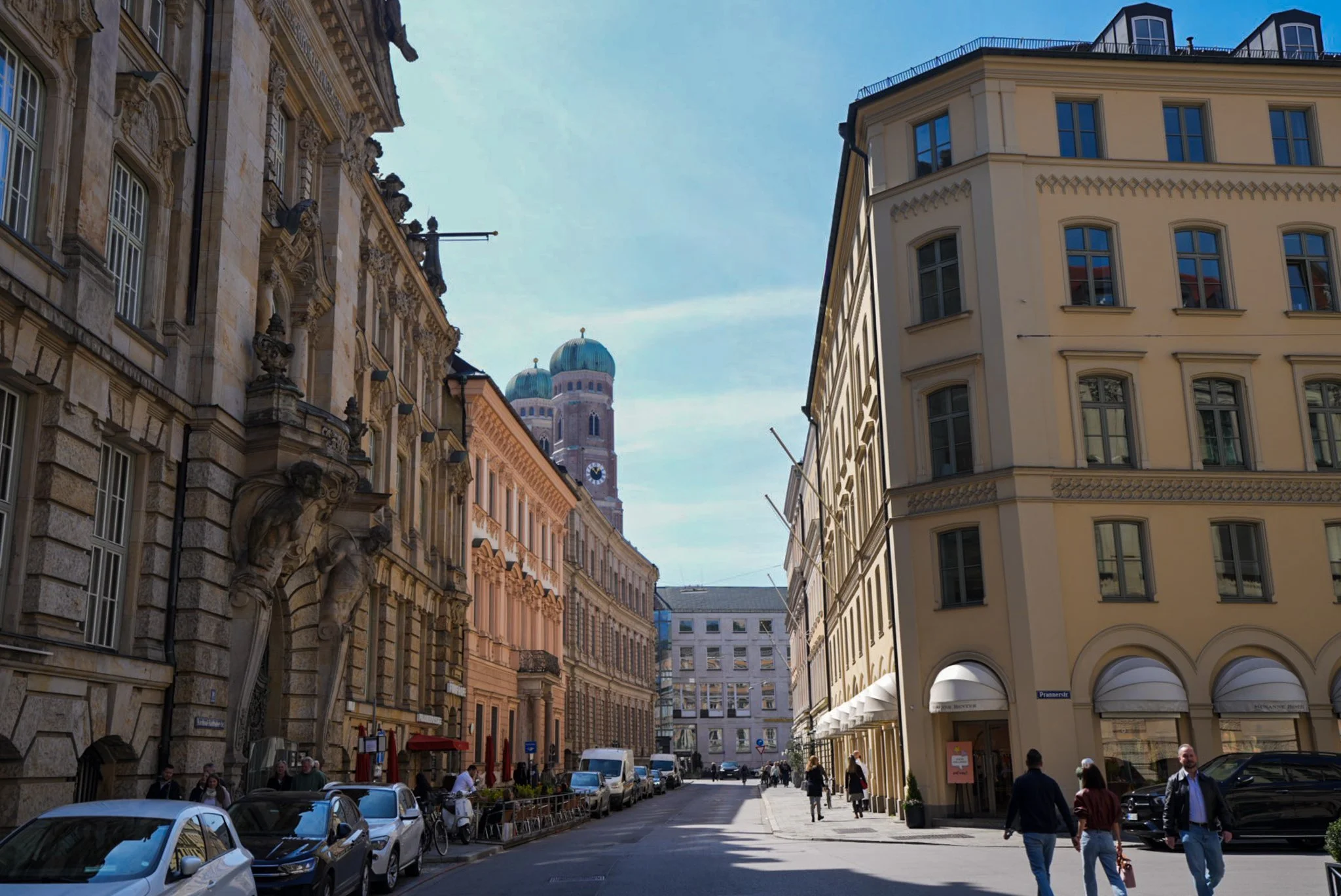 Straße in einer europäischen Stadt mit historischen und modernen Gebäuden, Menschen gehen, Autos parken, Himmel ist klar.