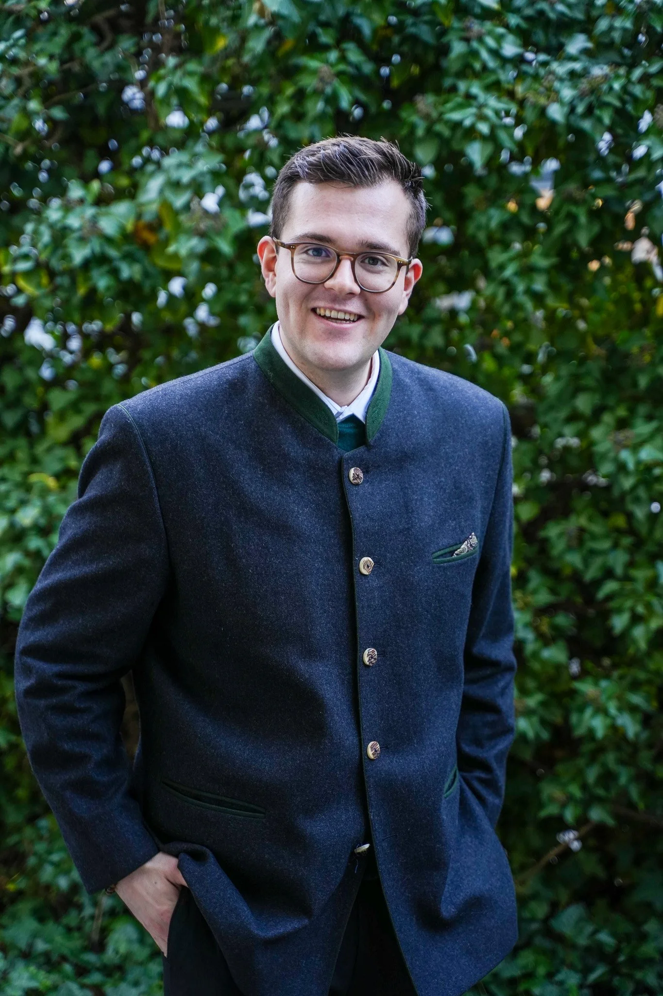 Junge Mann in traditionellen bayerischen Tracht, die vor einem grünen Laubstrauch steht.