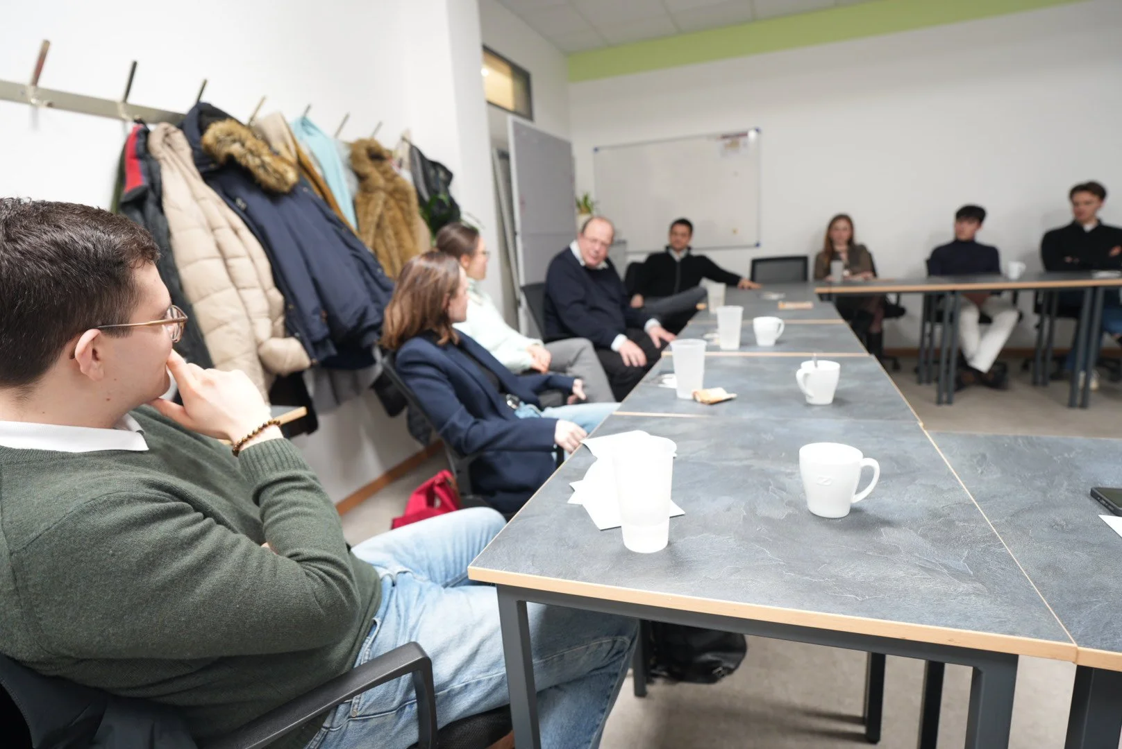 Menschen in einem Meetingraum sitzen an einem länglichen Tisch, einige sprechen, andere hören zu, in der Ecke hängen Mäntel an einer Garderobe.