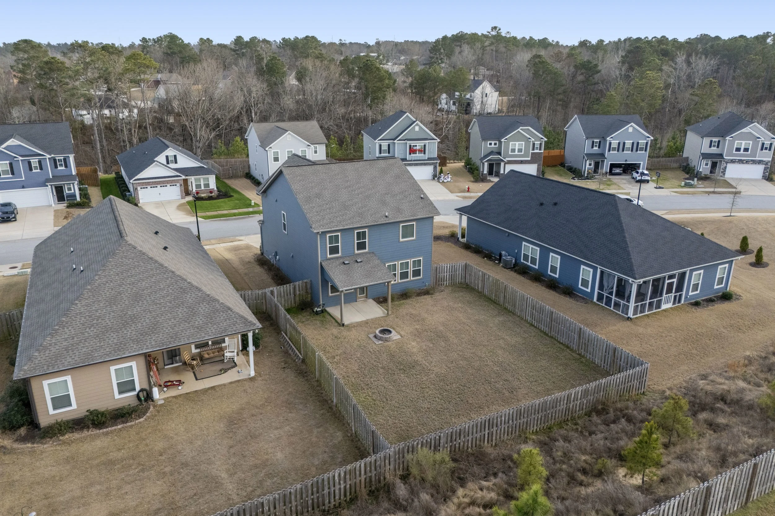 Aerial view of a suburban neighborhood featuring several houses with a fenced backyard, a fire pit, and a lawn, with additional houses and trees in the background.