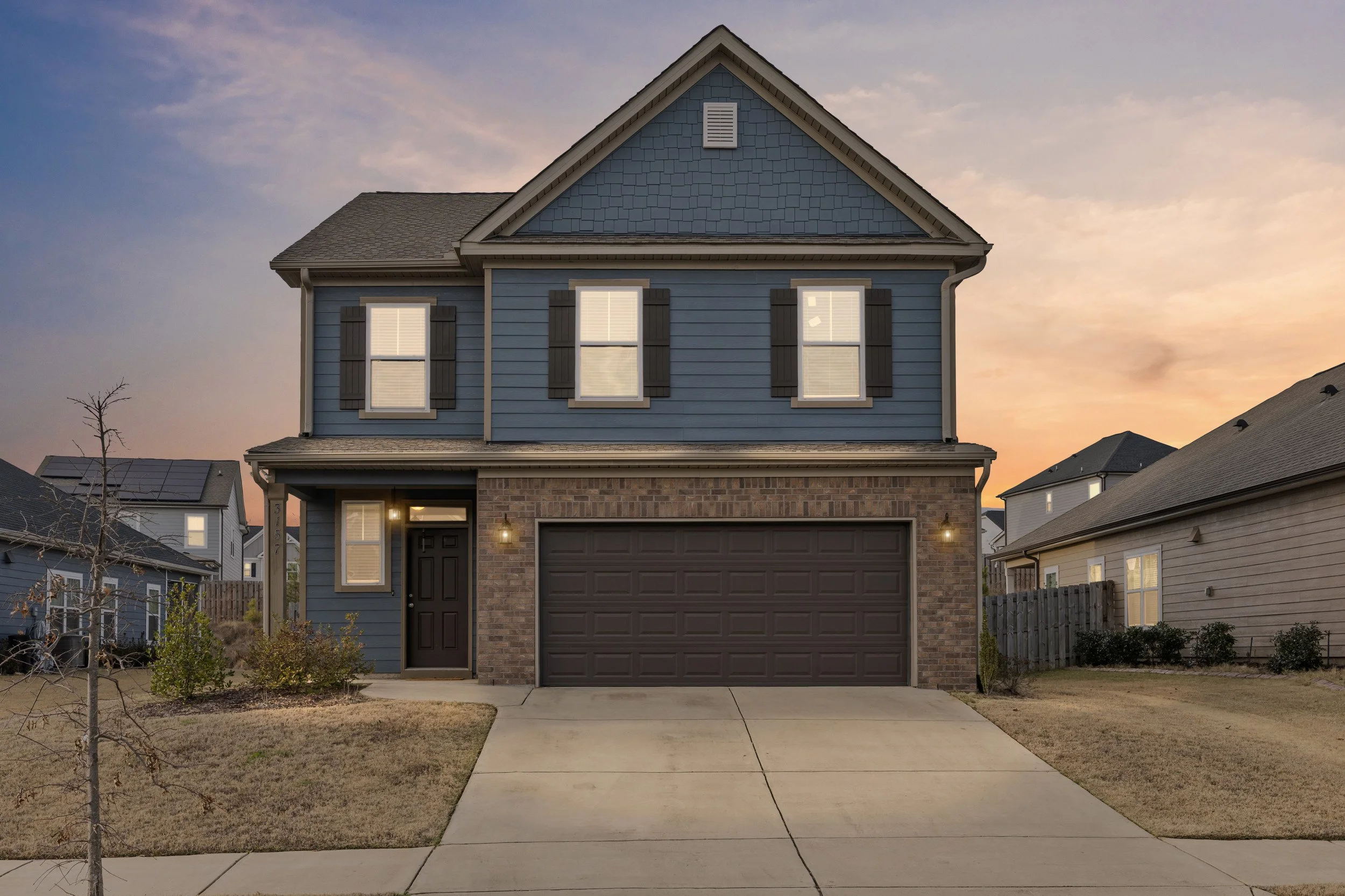 A two-story house with blue siding, black shutters, and brick accents, situated in a suburban neighborhood at sunset. The house has a dark garage door and a front door with a window, along with a small front yard and driveway.