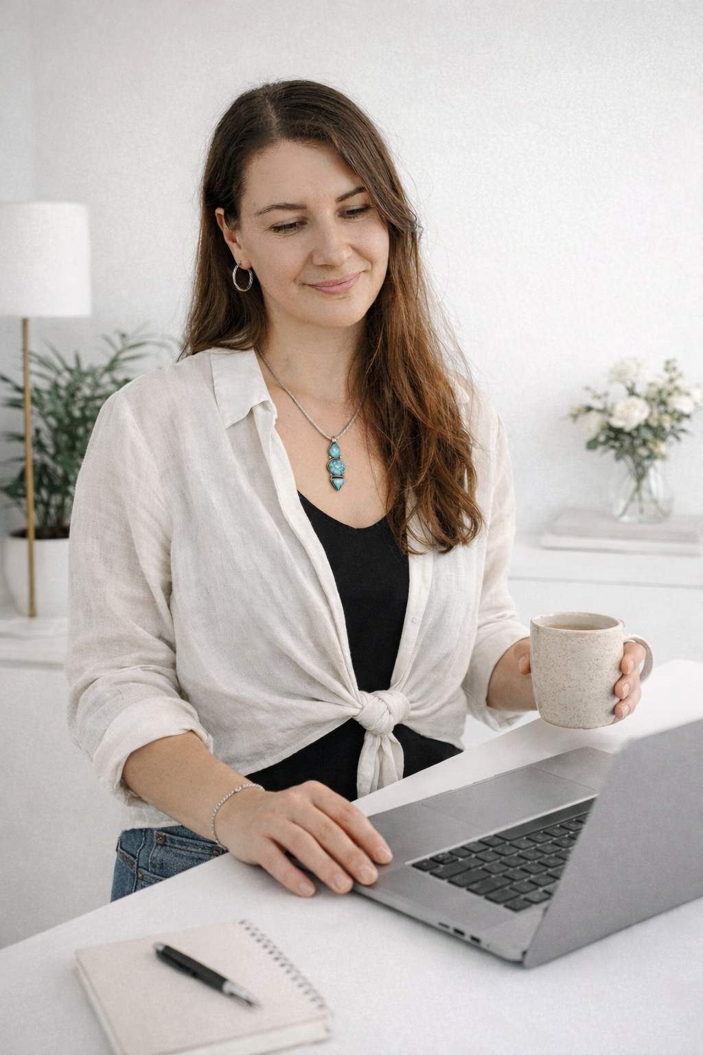A woman with long brown hair, wearing a white blouse tied at the waist, a black top, and jewelry, using a silver laptop while holding a coffee mug in a bright, minimalistic room with a notebook and pen on the table.