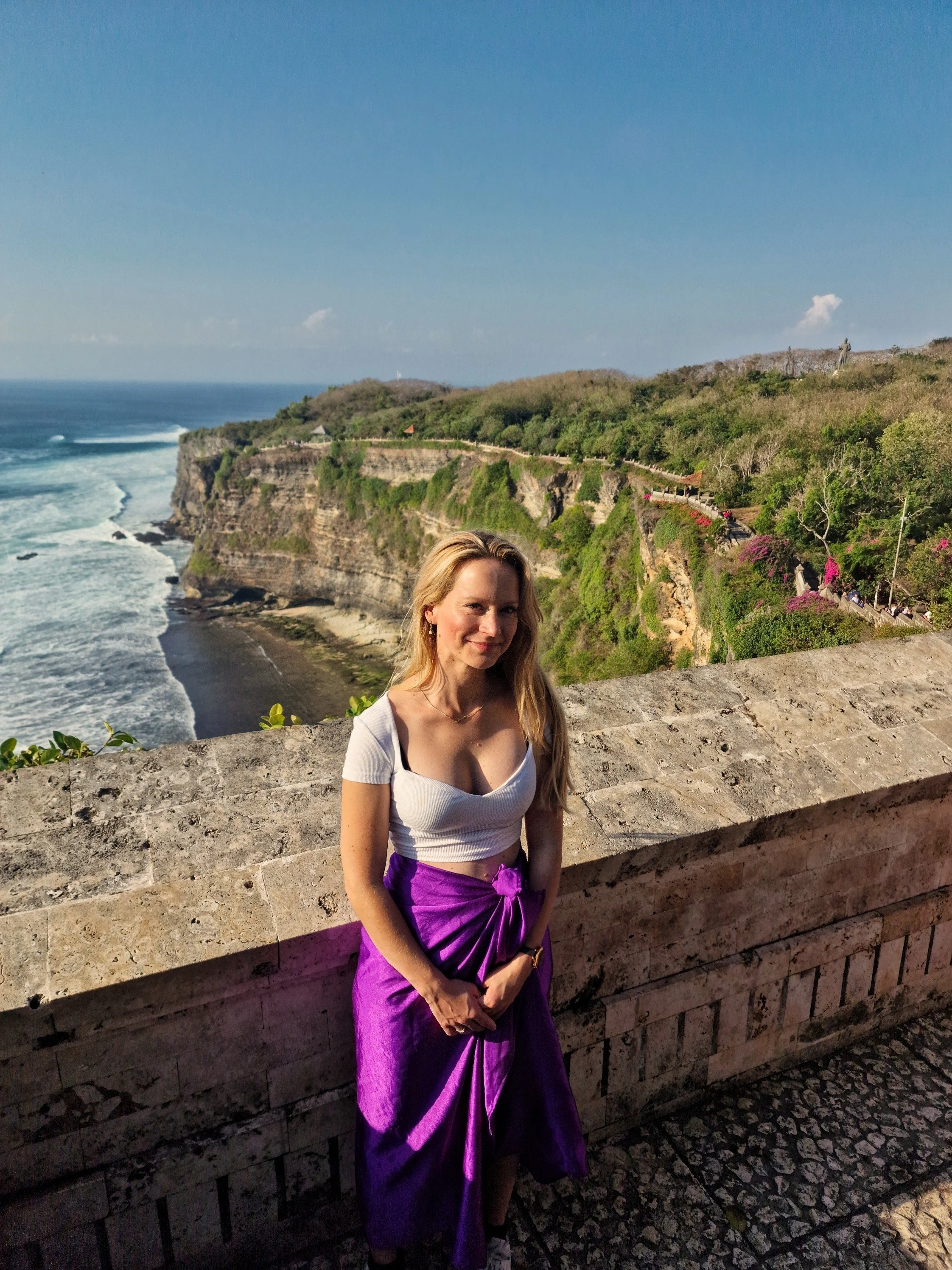 Een vrouw met lang blond haar staat tegen een stenen muur met een kustlijn met kliffen en de oceaan op de achtergrond.