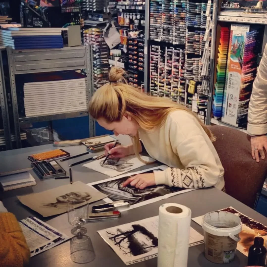 Vrouw die in een boetiek houtskooltekeningen of schetsen maakt, met kunstbenodigdheden en foto's op tafel, omringd door kleurpotloden, stiften en kunstboeken.