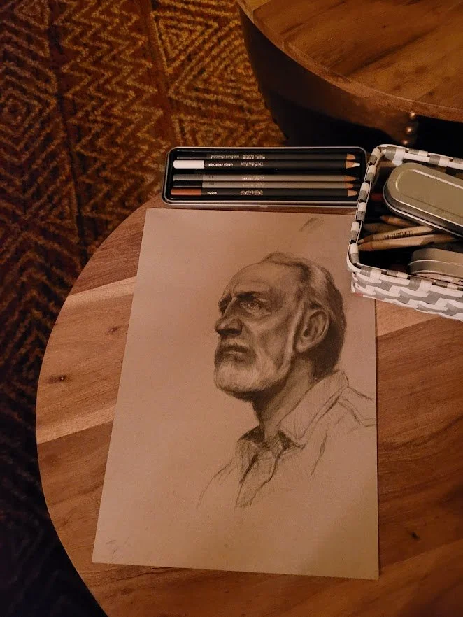 Potloodschets van een oudere man met een baard en korte haren, op een houten tafel met tekenpotloden, doosjes en een stalen blikje.