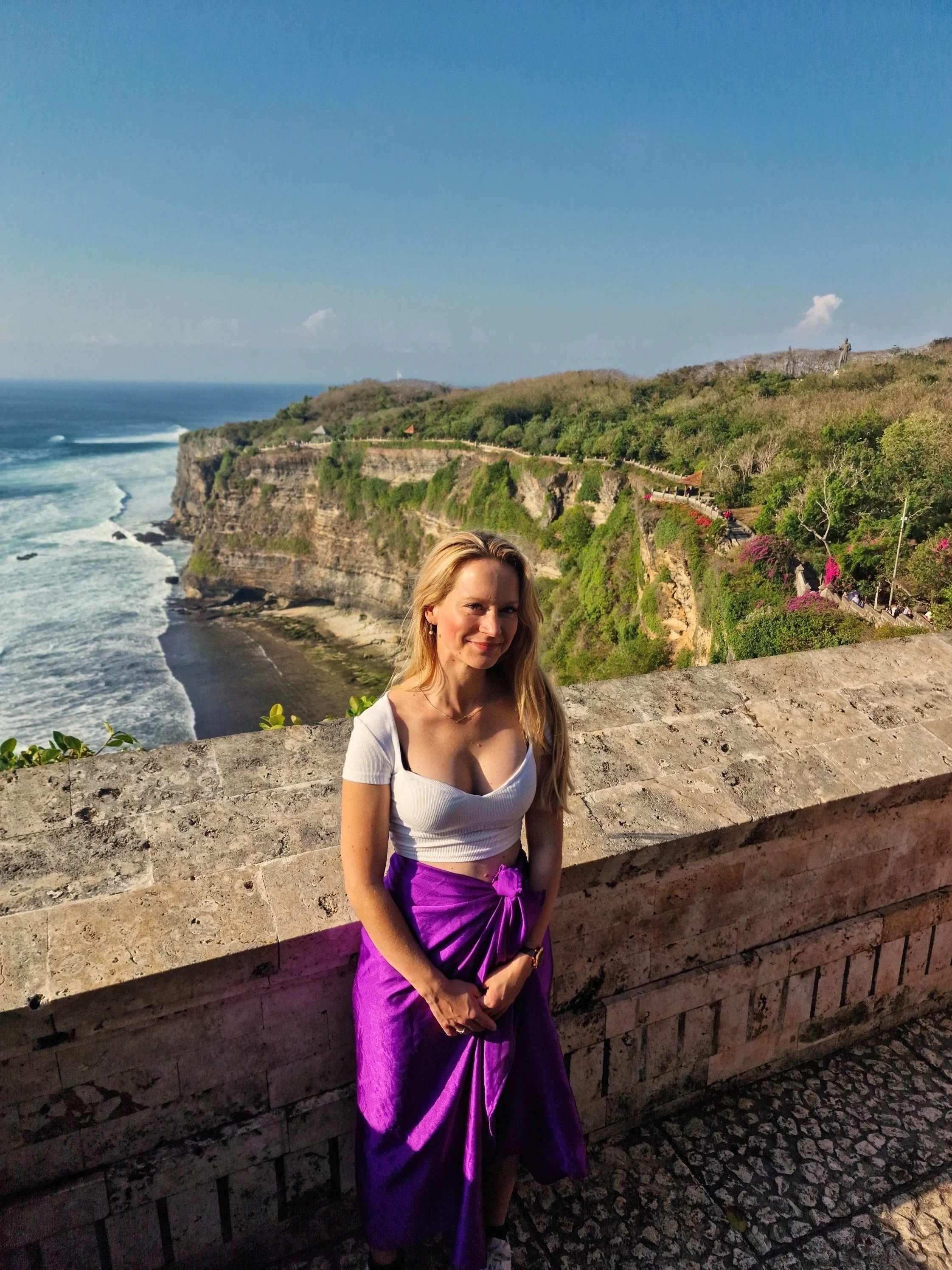 Een vrouw staat voor een bakstenen muur met uitzicht op een klif en de oceaan op de achtergrond.