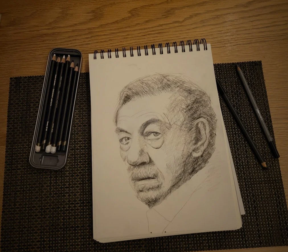 Potloodschets van een man op een schetsboek met potloden en een pen op een houten tafel.