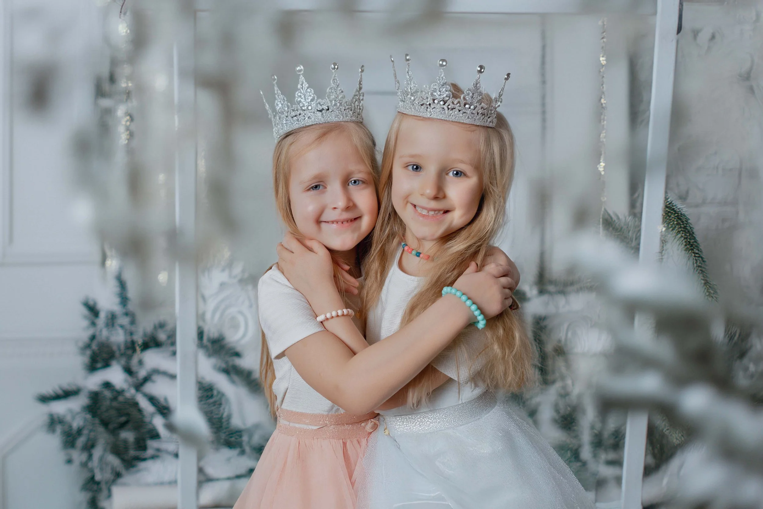 Deux jeunes filles avec des couronnes brillantes, s'embrassant et souriant dans un décor de Noël avec des sapins enneigés en fond.