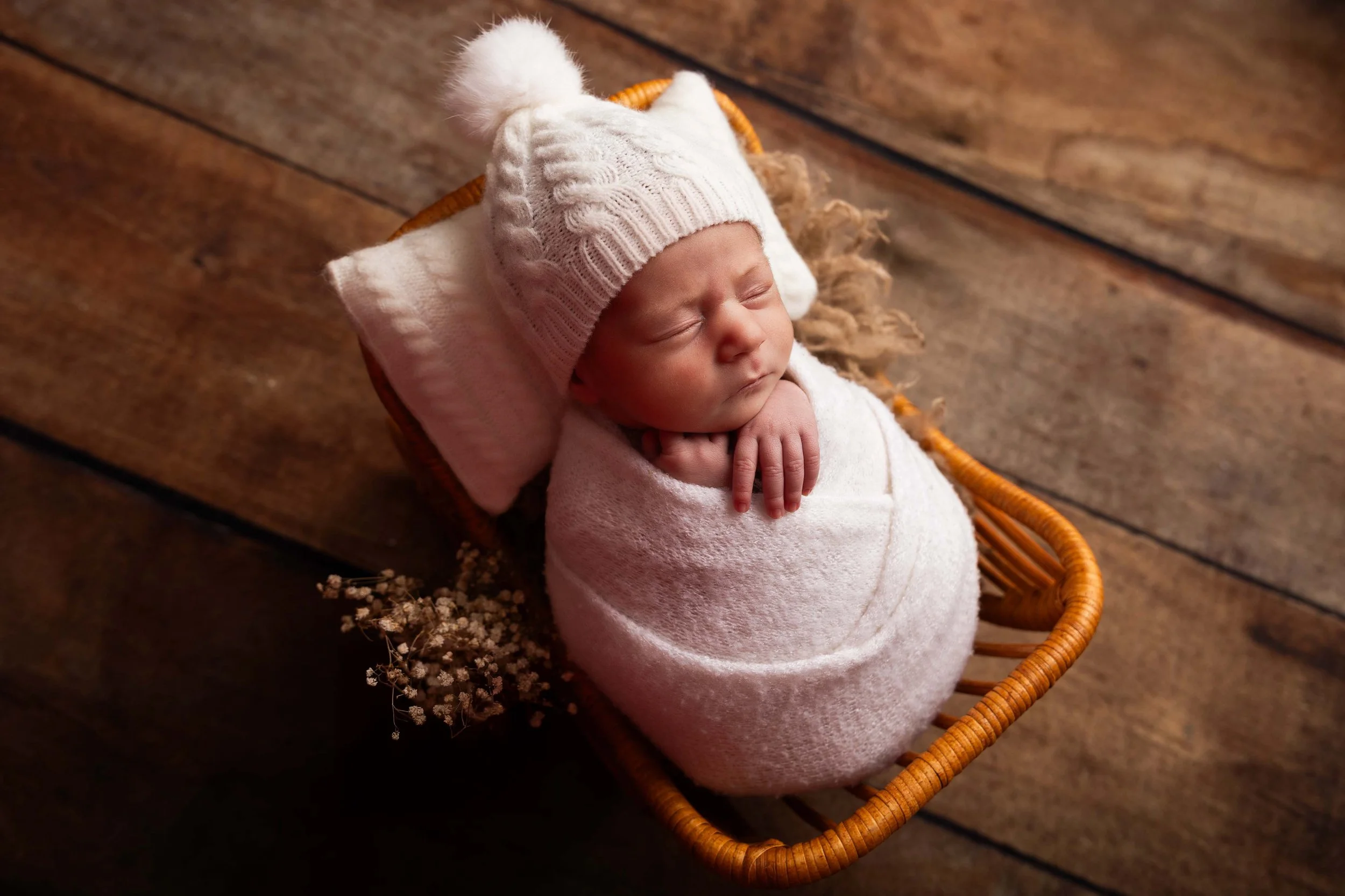 Bébé endormi dans un lit en osier, portant un bonnet en laine blanc avec des oreilles et enveloppé dans une couverture blanche, sur un sol en bois.