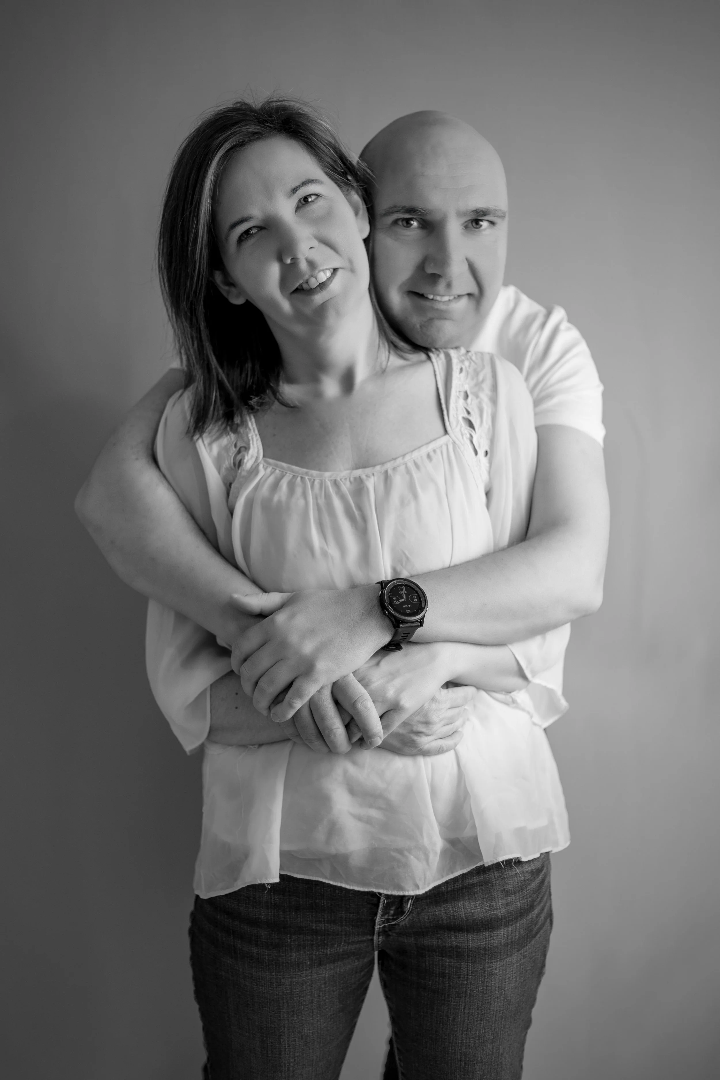 Une femme et un homme posent ensemble, la femme est devant et l'homme l'enlace derrière, eux souriant, dans un style monochrome.