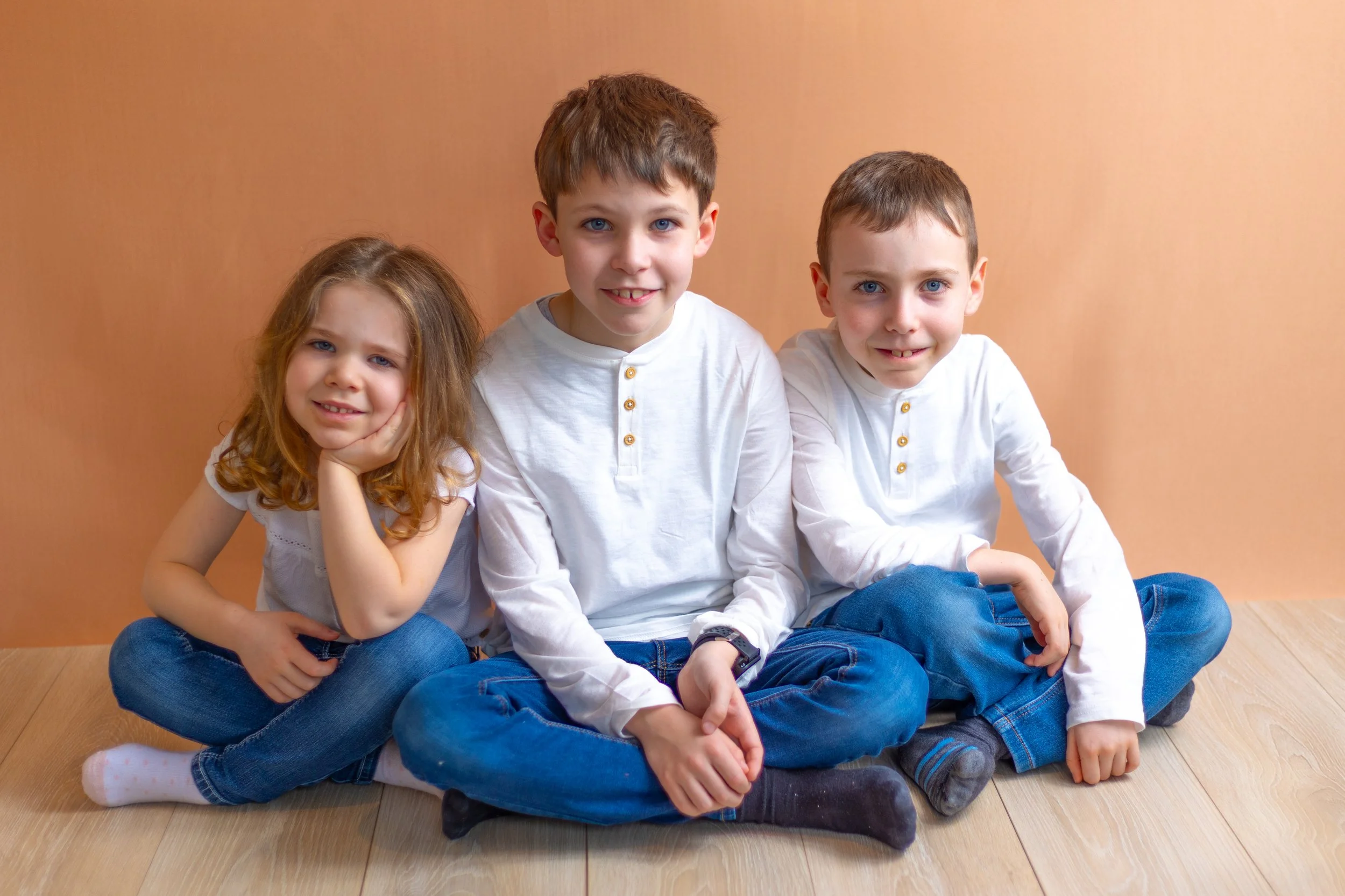 Trois enfants assis sur le sol en bois devant un mur couleur pêche, deux garçons et une fille, tous portant des vêtements décontractés blancs et jeans, souriants.