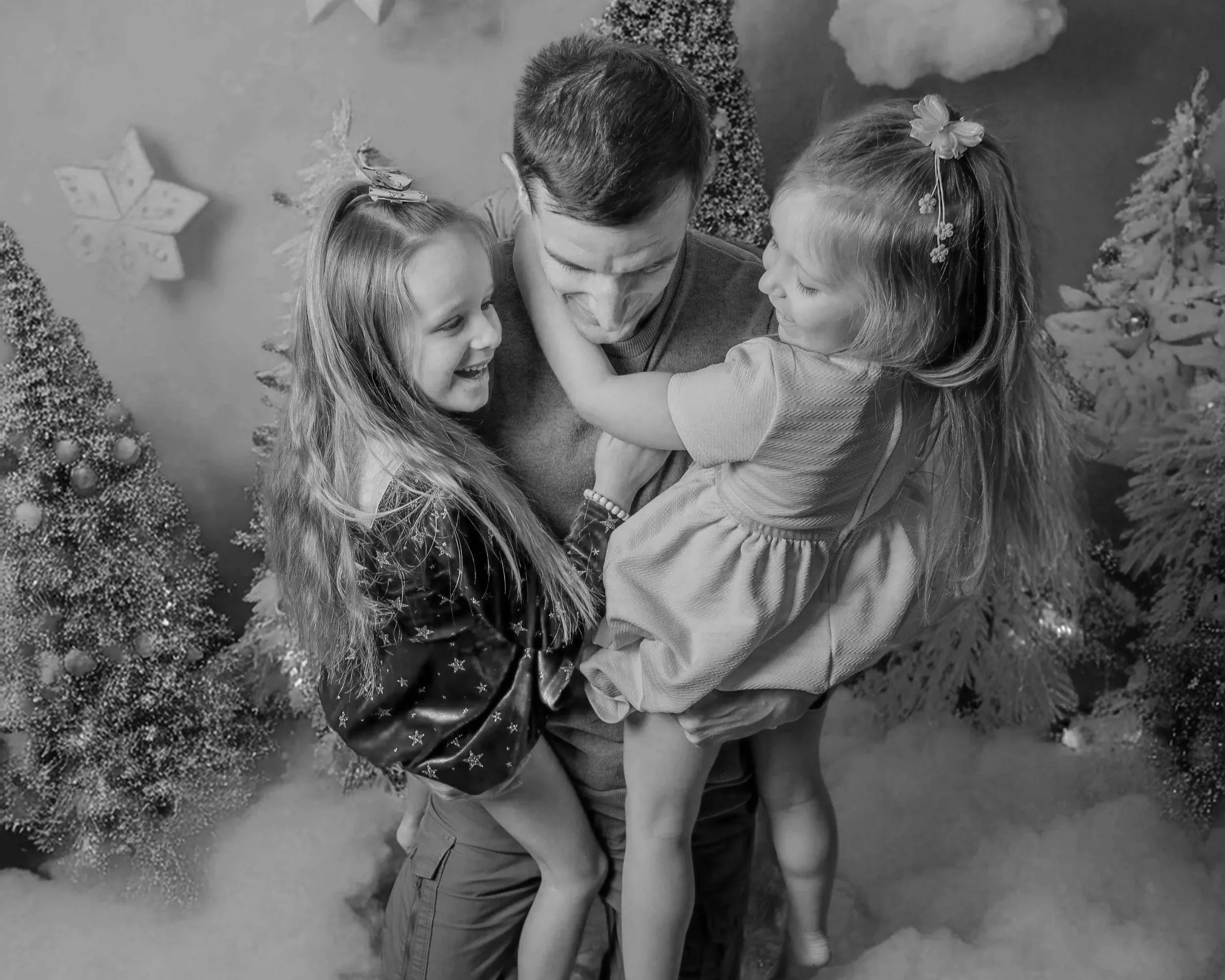 Un homme tenant deux jeunes filles dans un décor de Noël avec des arbres et des flocons en arrière-plan, tous souriants et riant.