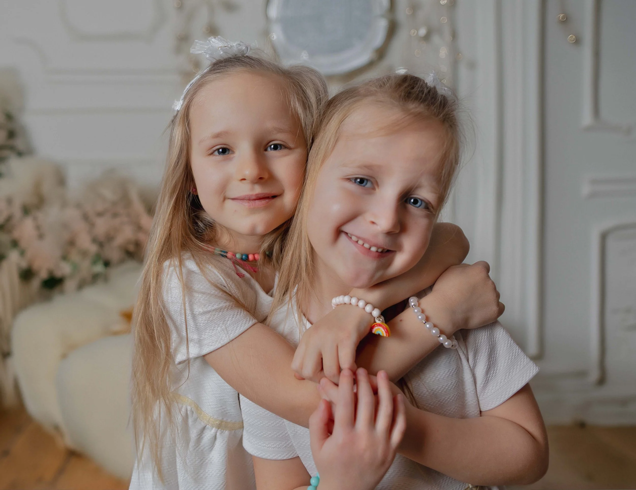 Deux jeunes filles soulagées, souriantes, se tenant l'une l'autre dans une pièce lumineuse avec des décorations florales en arrière-plan, portant des vêtements blancs et des accessoires colorés.