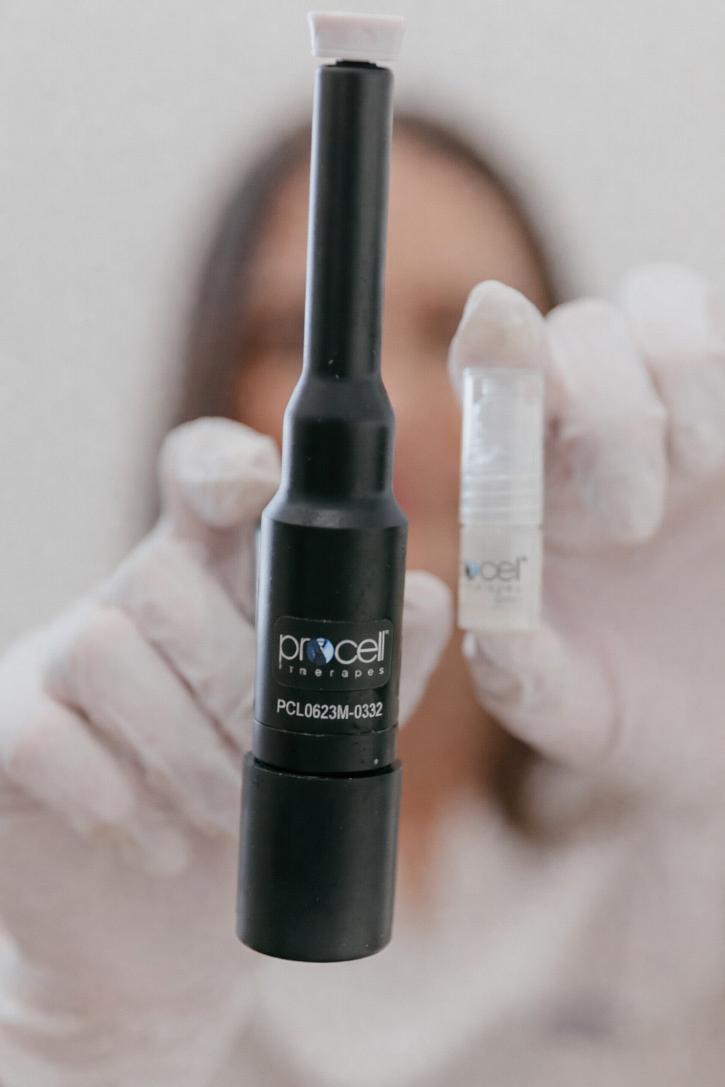 Close-up of a medical professional holding a black Procell Therapies device, with a small container in the background, blurred face and white gloves visible.