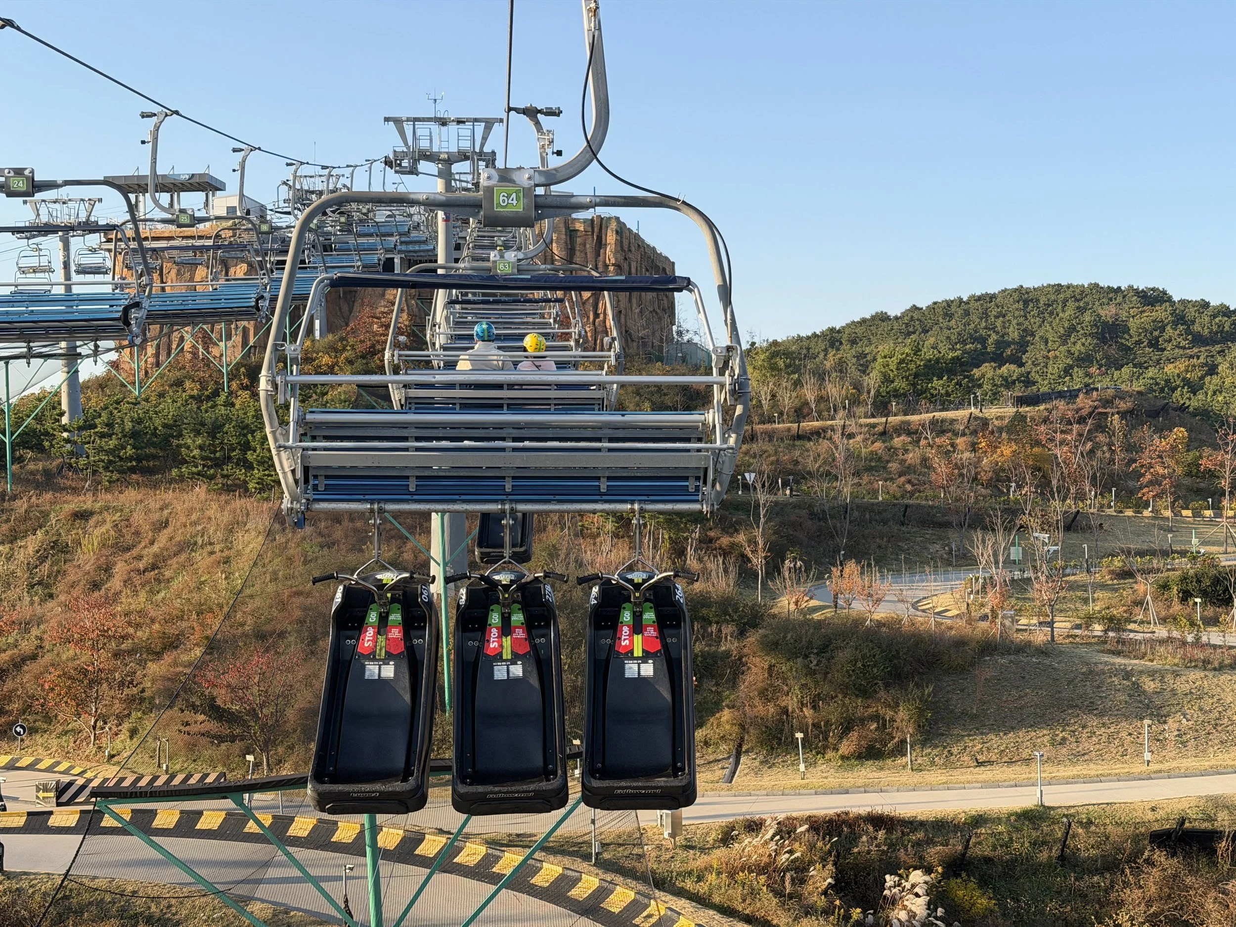 A ski lift with two empty seats carrying two children with helmets up a mountain, with trees and clear blue sky in the background.