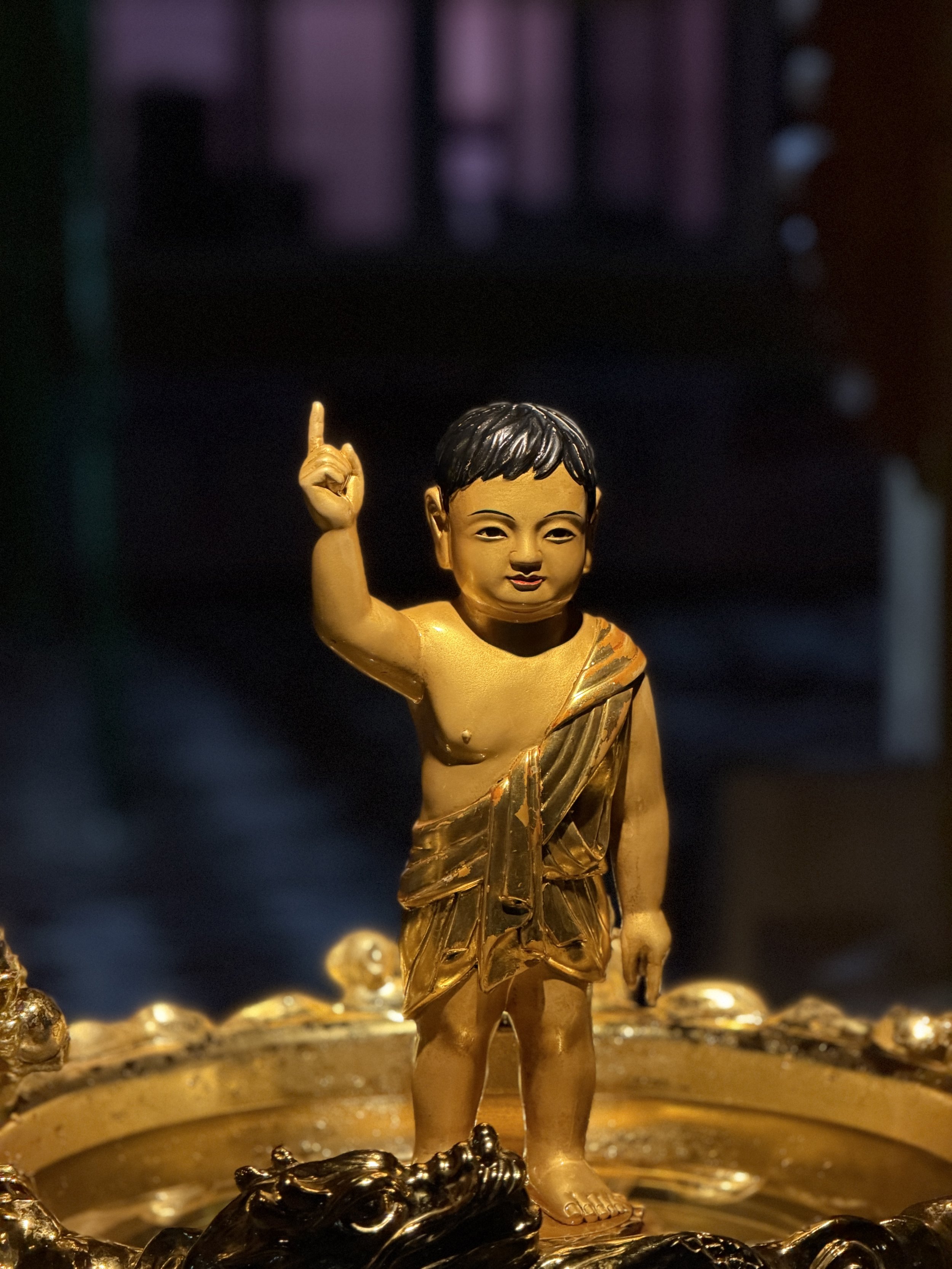 A close-up of a gold-colored figurine of a young boy with short hair, raising his right index finger, standing on a decorative base with a water feature.