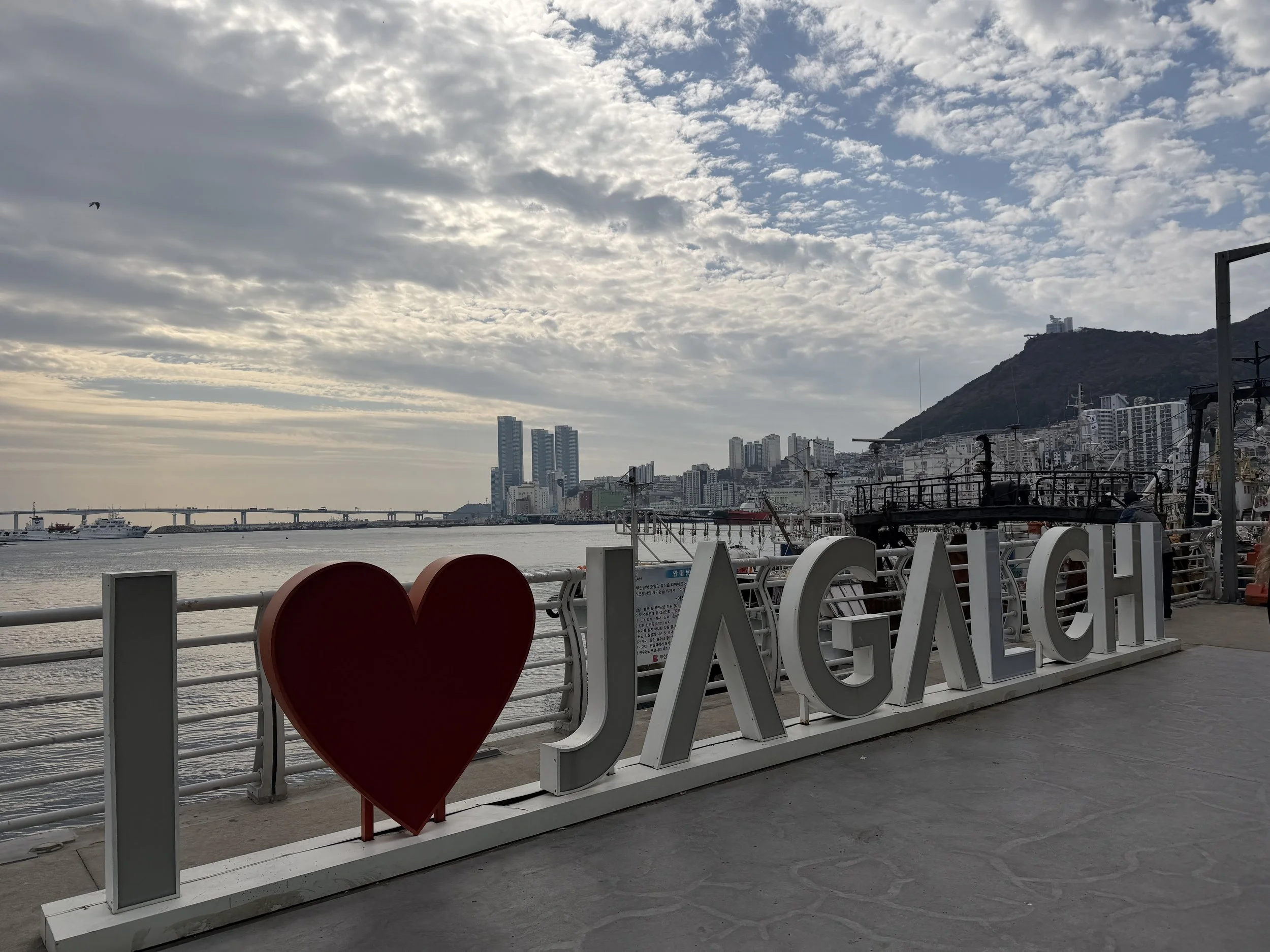 A waterfront scene with a skyline of tall buildings, a harbor with boats, and a mountain in the background. There is a decorative installation with the words 'I LOVE U' on a railing in the foreground.