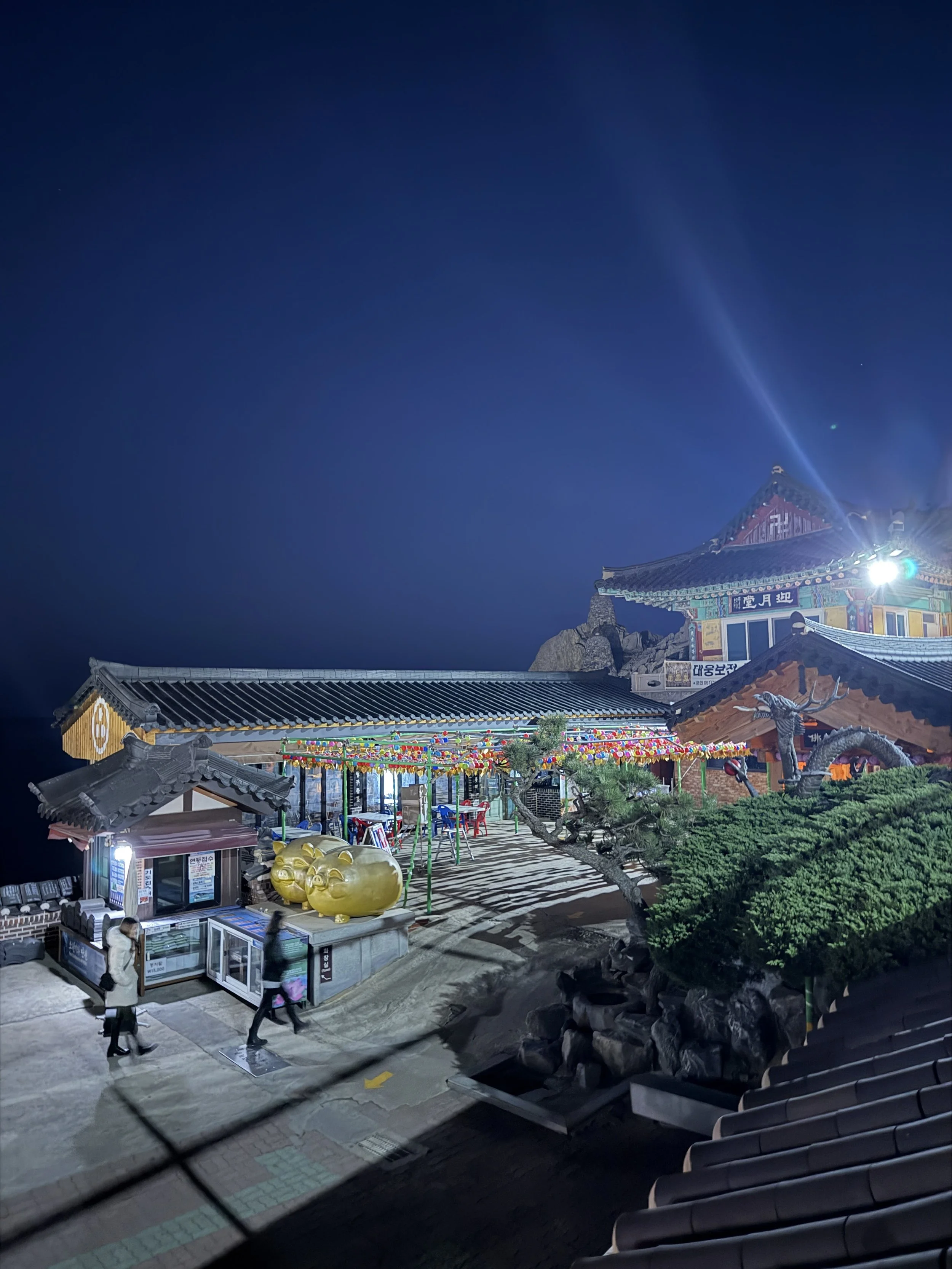 Night view of a traditional Korean temple with colorful roof and intricate architecture, illuminated with lights. The courtyard has a small amusement ride with colorful decorations, large golden pig statue, and a few people walking around. The sky is