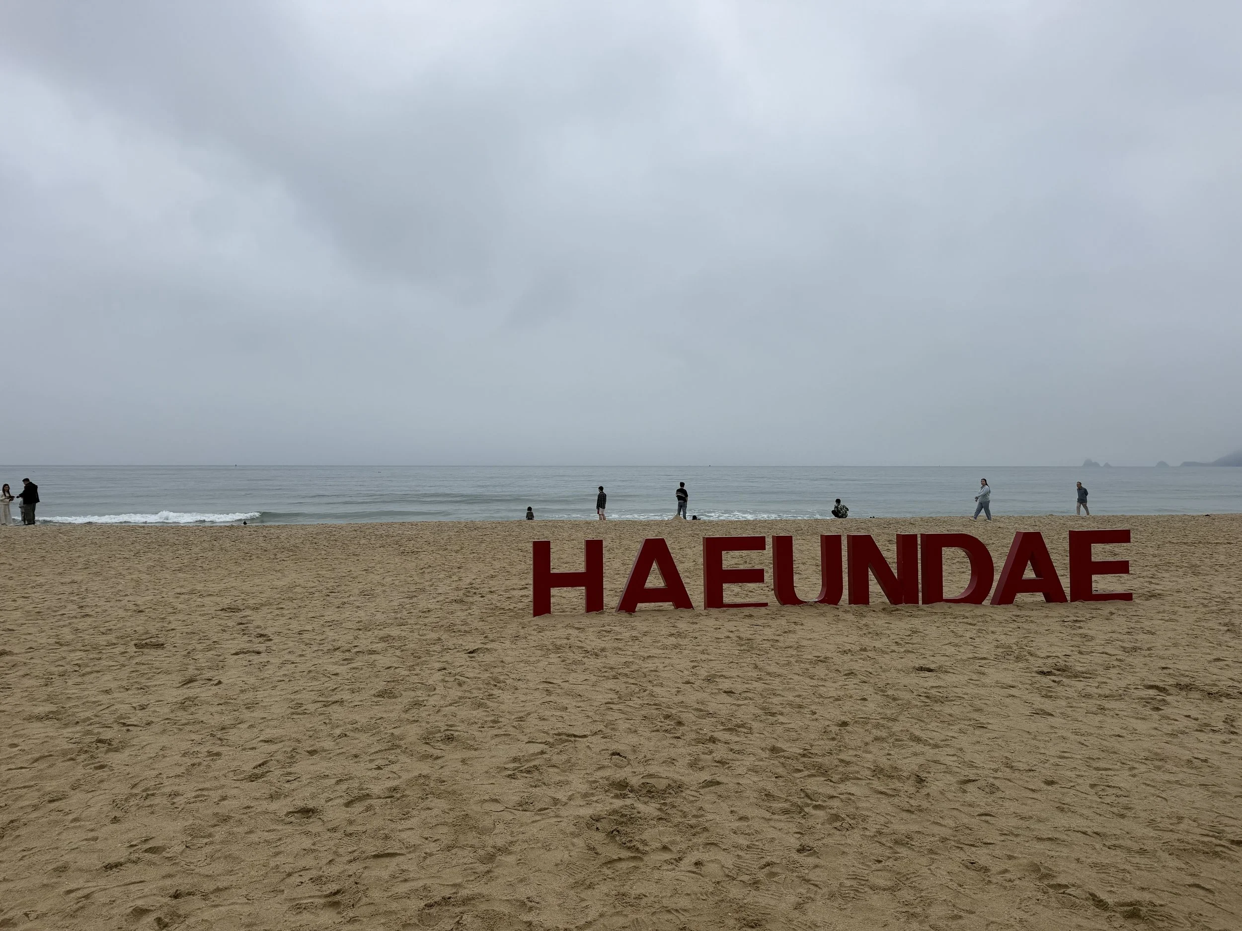 Cloudy day at the beach with people walking along the shore, and a large red sign with partially visible text in front of the sand.