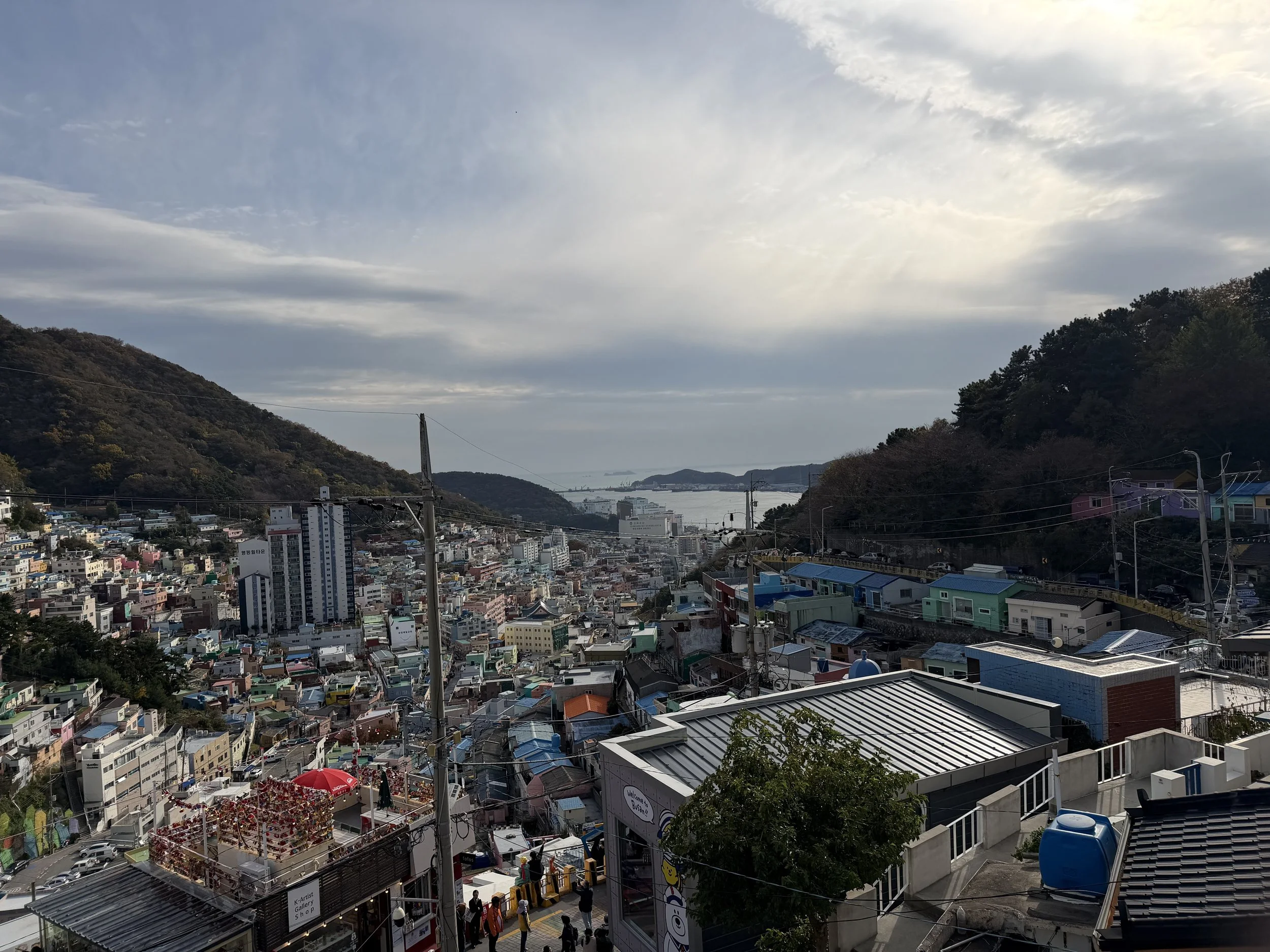 A view of a city nestled between hills with a body of water in the distance under a cloudy sky.