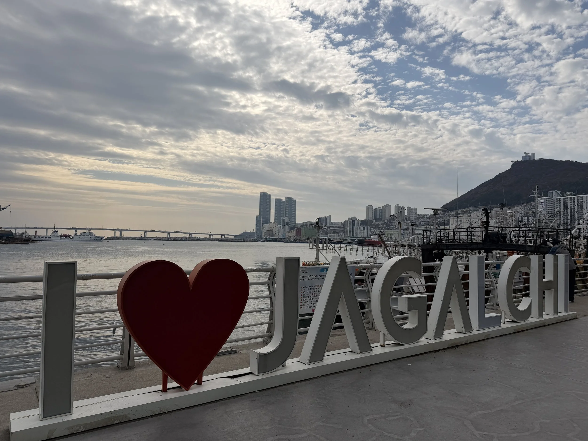 The image shows a waterfront with a large white sign that spells out 'I ❤️ SEO.' The sign is on a board on a promenade, with a cityscape and a hill with buildings and a communication tower in the background. The sky is cloudy, and there are ships doc