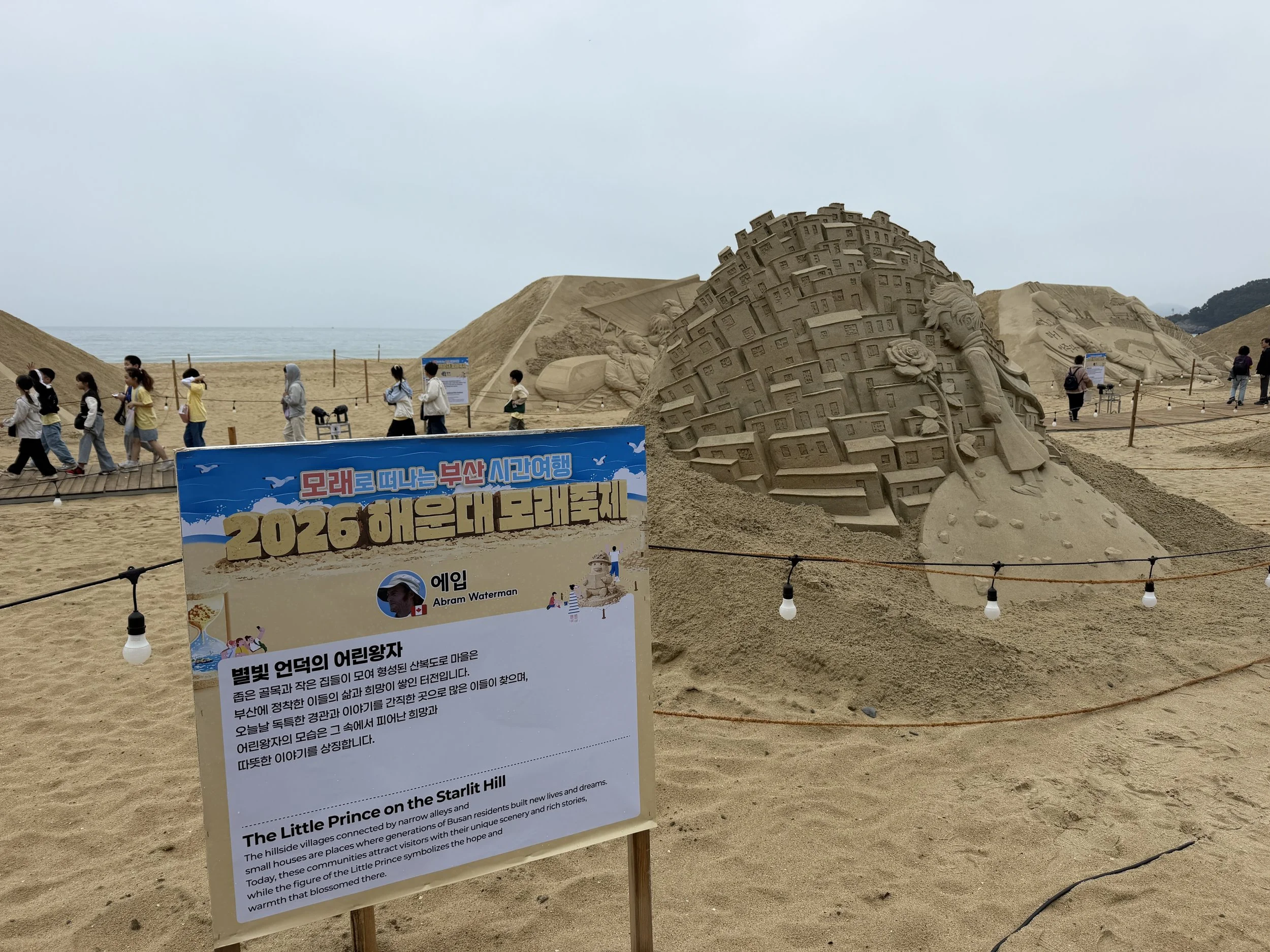 Sandcastle sculptures on the beach depicting a woman with flowing hair, a small house, and other figures. People are walking past the sculptures, and informational signs are displayed in front of the sand art.