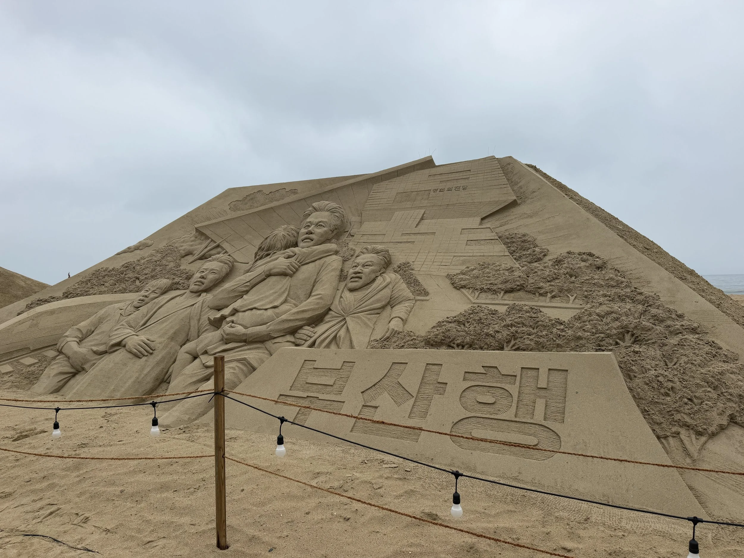 Sand sculpture of the iconic Mount Rushmore with faces of U.S. presidents carved into the sand, under a cloudy sky, surrounded by a wooden barrier with hanging lights.
