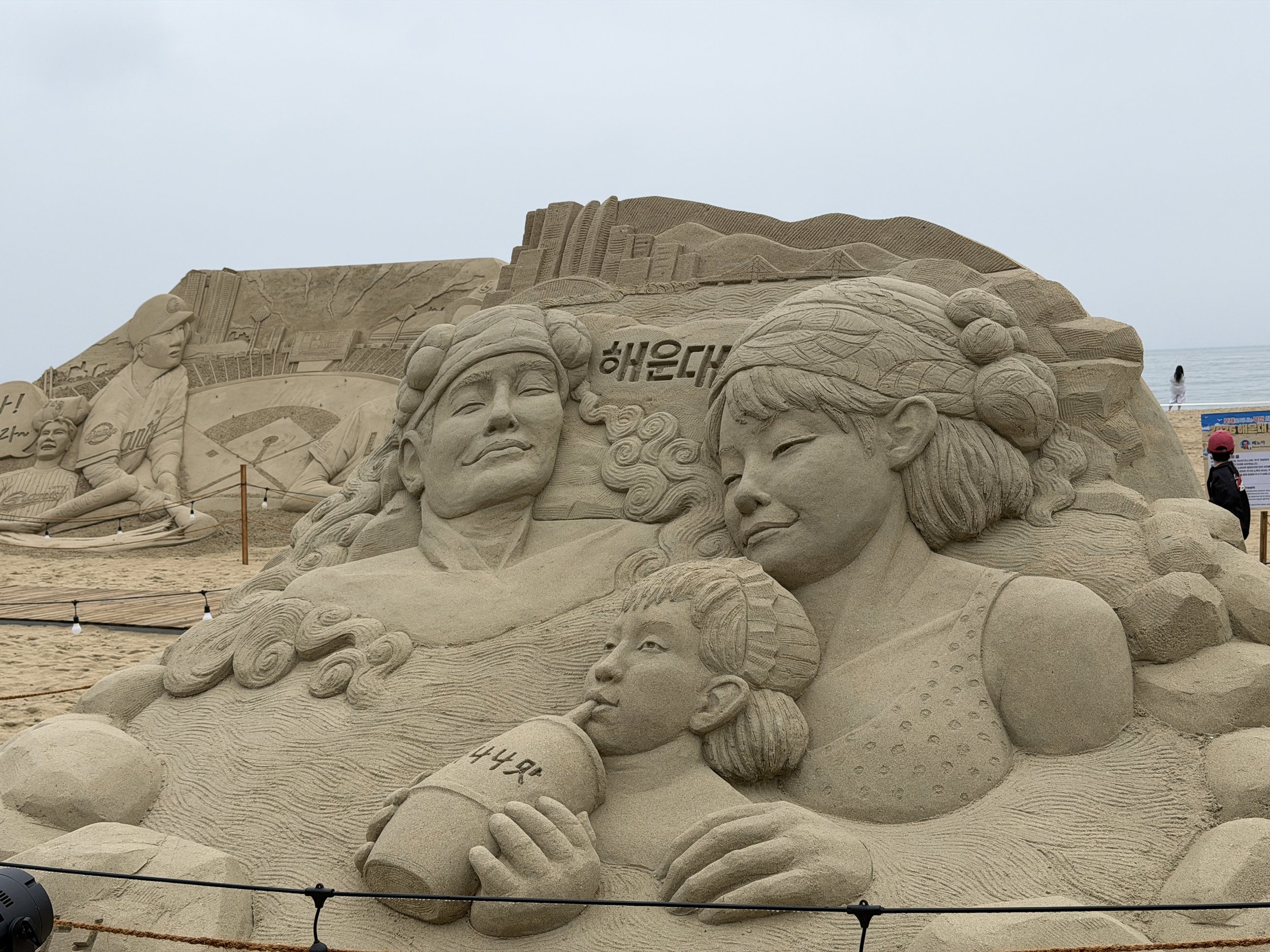 A large detailed sand sculpture on a beach depicting various figures and scenes, including a woman with a headscarf, a woman with glasses, and a monument with Korean characters. The sculpture features intricate carvings of mountains, bridges, and oth