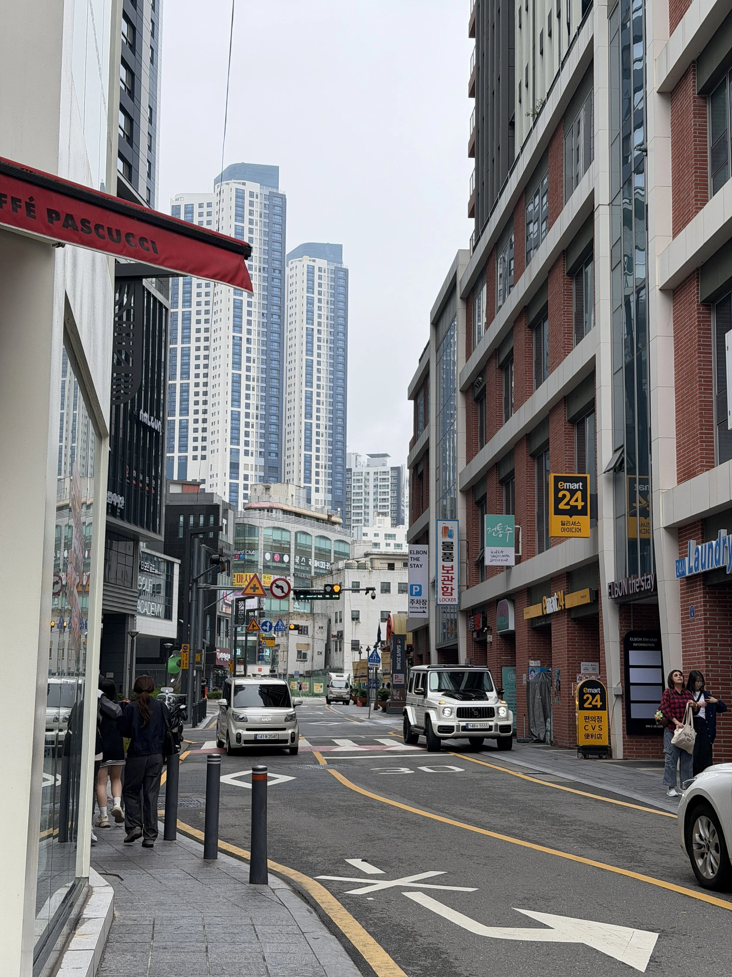 City street scene with tall modern buildings, cars, pedestrians, and various signs including a CVS pharmacy sign and traffic signals, under an overcast sky.