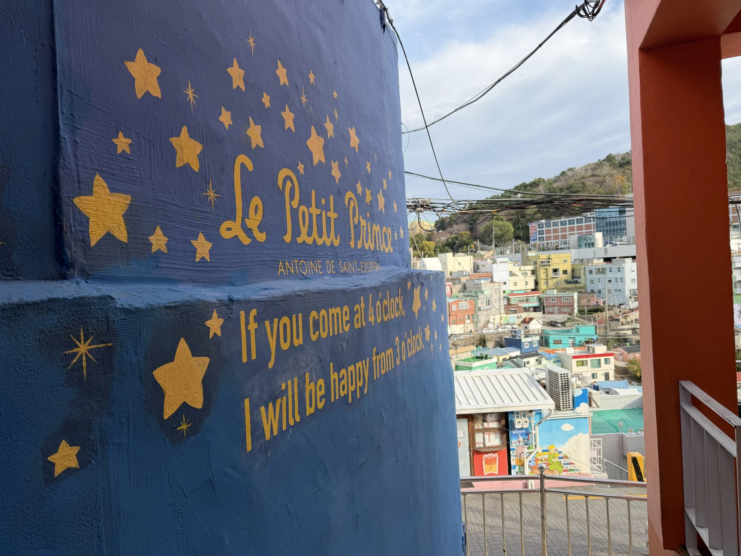 A colorful mural on a blue wall with yellow stars and the text 'Le Petit Prince' by Antoine de Saint-Exupery. In the background, there are buildings, hills, and a partly cloudy sky. Part of a red railing is visible on the right side of the image.