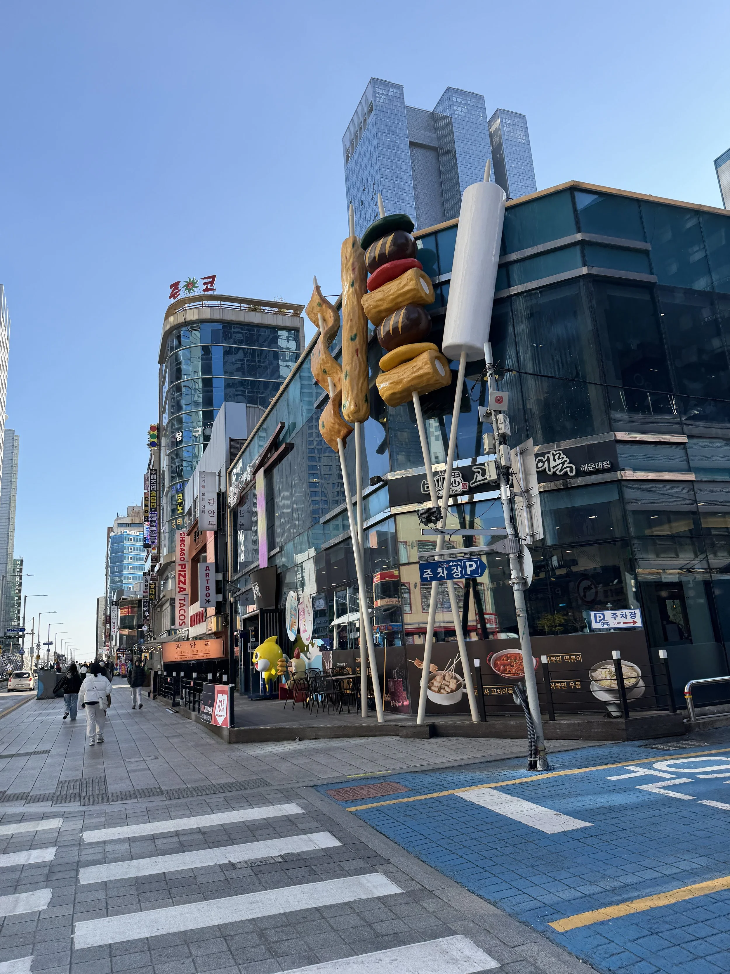A busy urban street scene with pedestrians, modern glass buildings, and large decorative food sculptures of skewered dishes and dumplings.